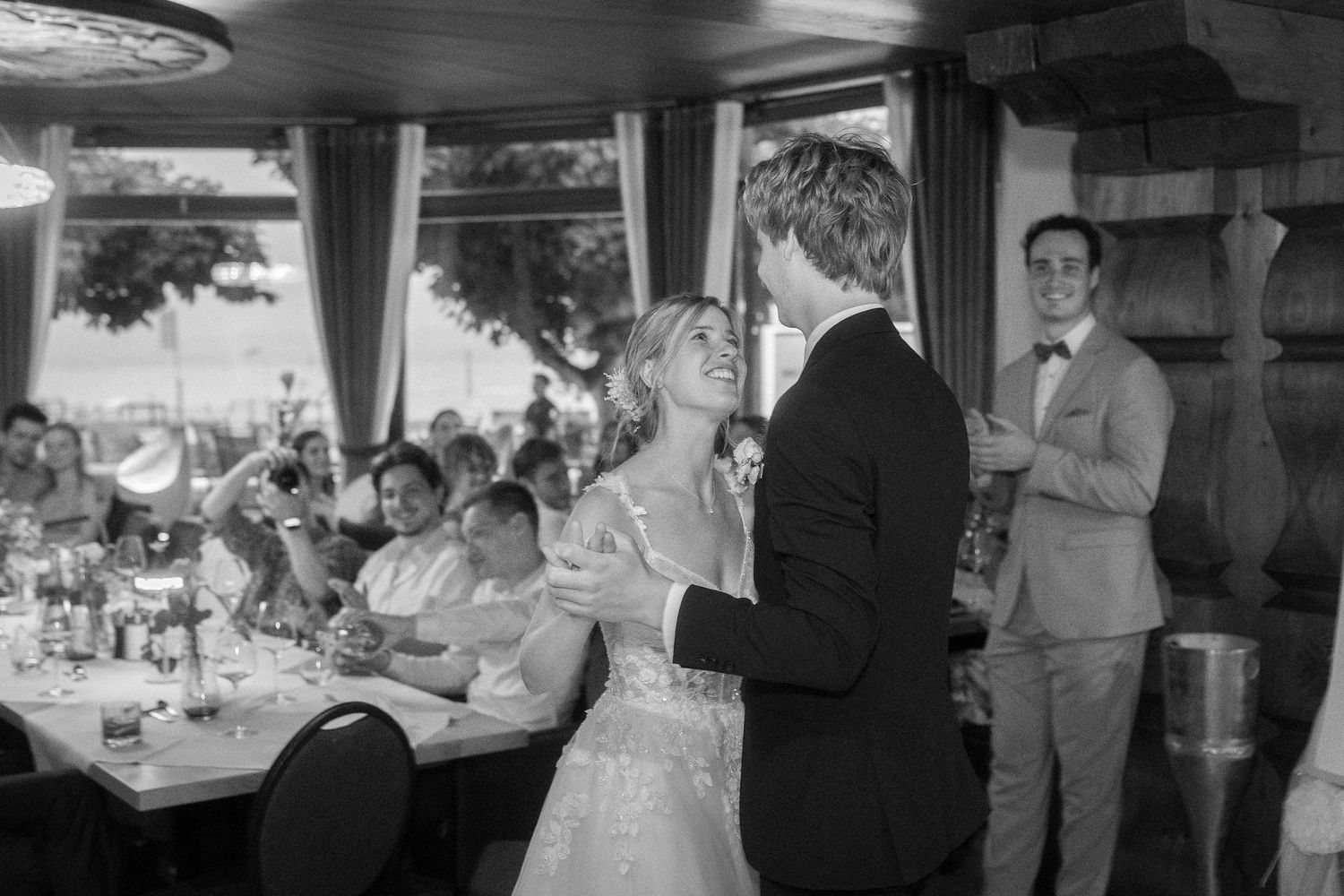 Bride and groom share a joyful first dance at a wedding reception while smiling guests watch from tables in this black and white photo.