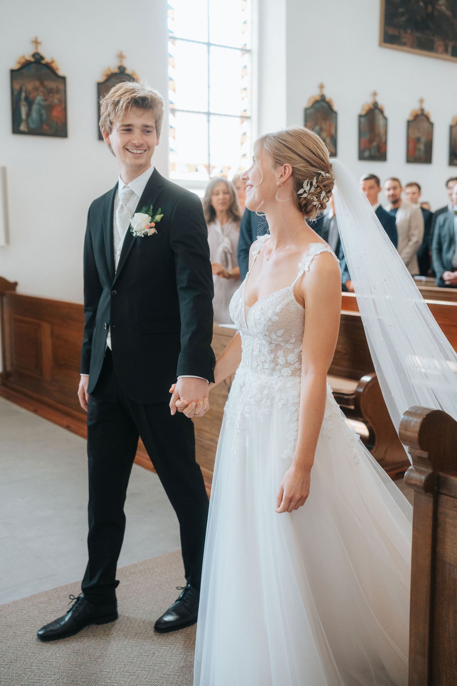 Smiling groom in black suit holds hands with bride in white lace gown at church altar.