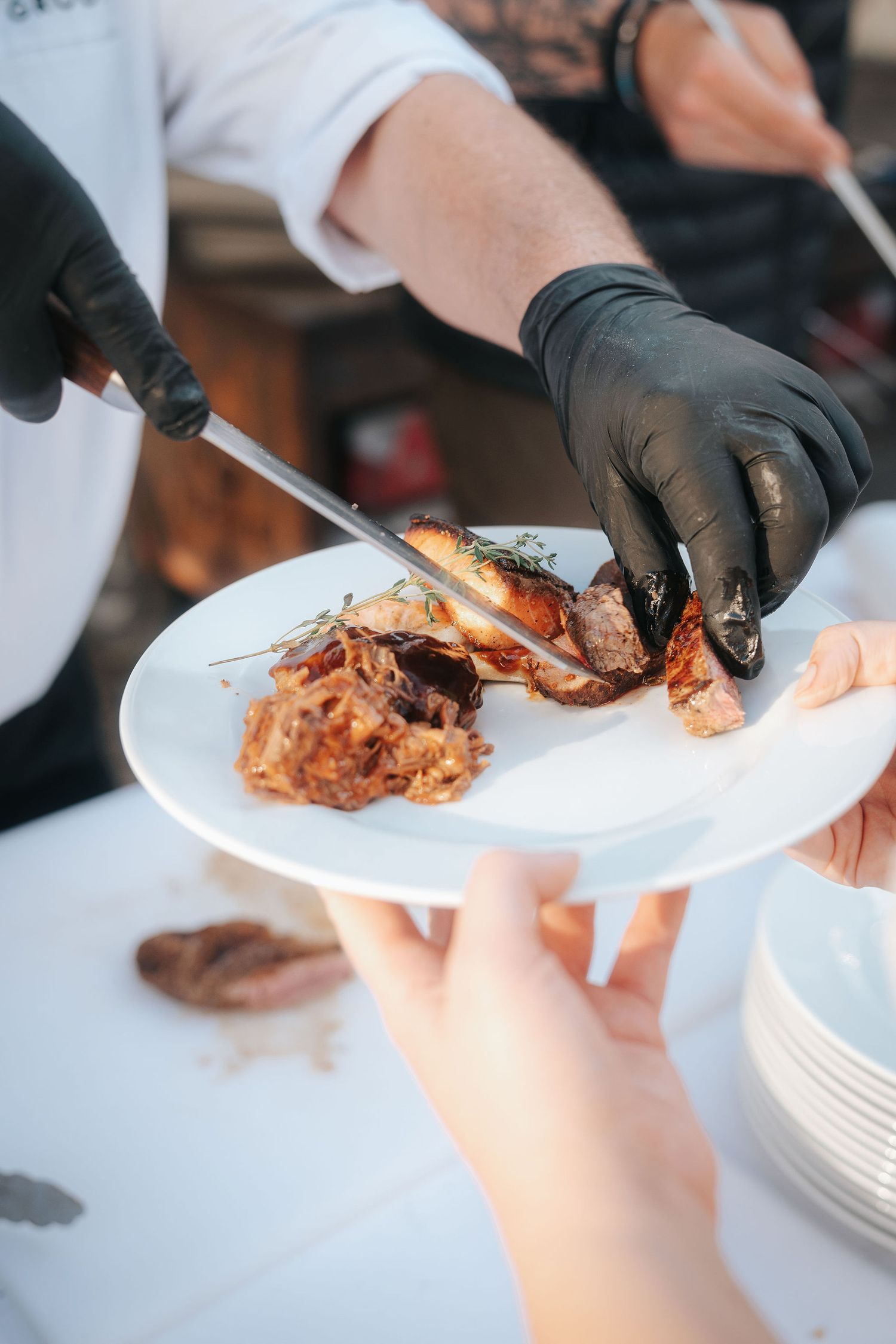 Chef in black gloves uses a knife to plate roasted meat with herbs on a white plate while a guest holds out a hand.