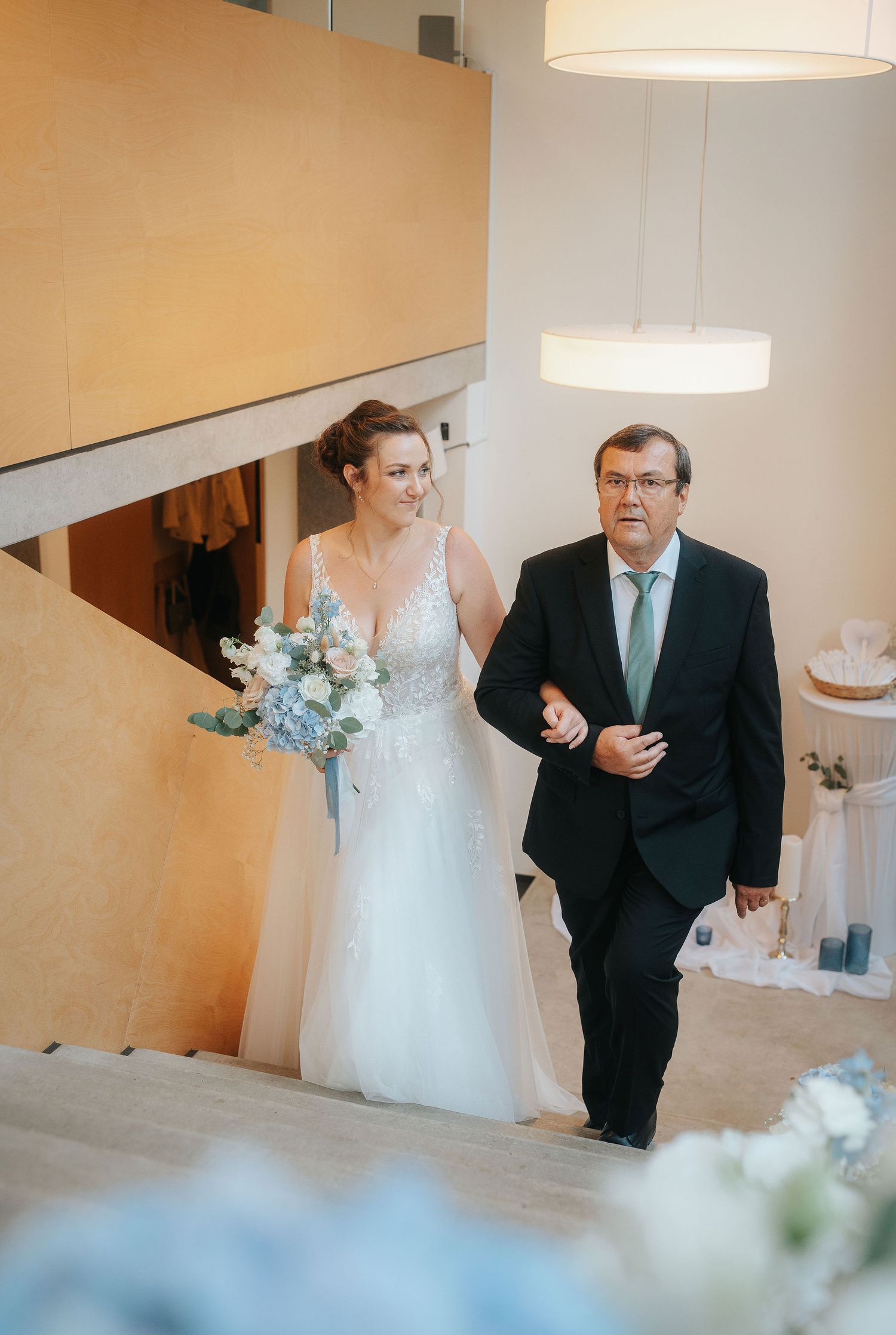A bride in a lace gown holding a blue and white bouquet walks up wooden stairs arm in arm with a man in a dark suit.