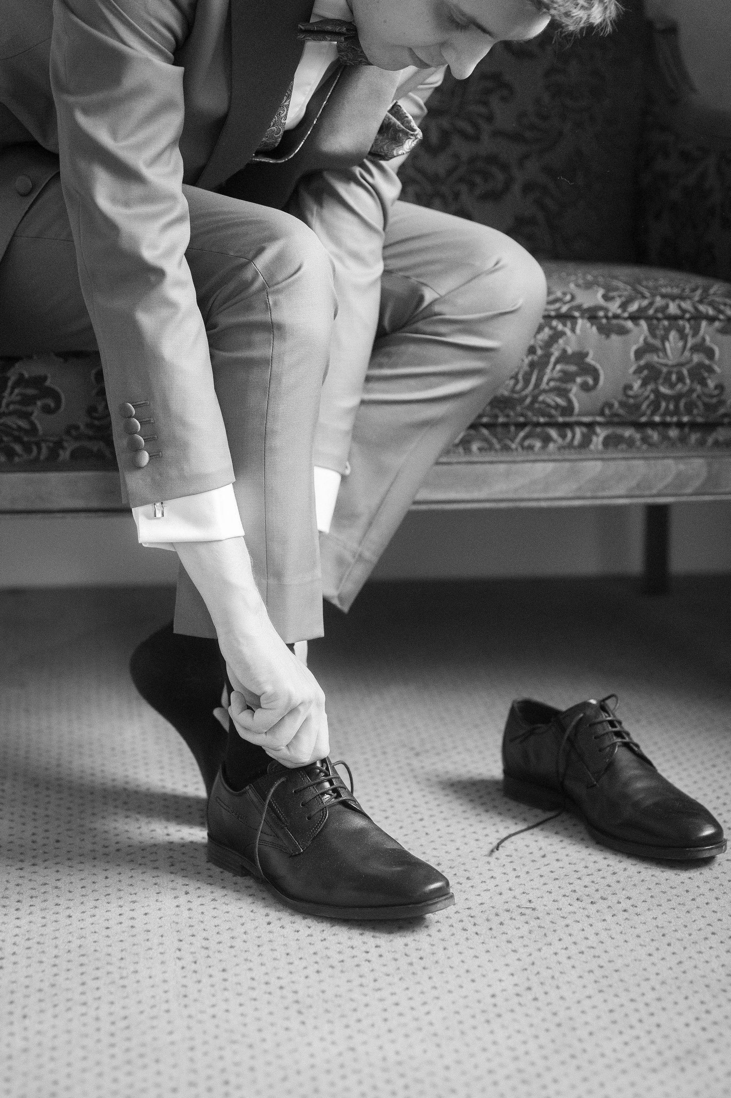 Black and white photo of a groom in a suit sitting on a vintage sofa while tying the laces of his black leather dress shoe.