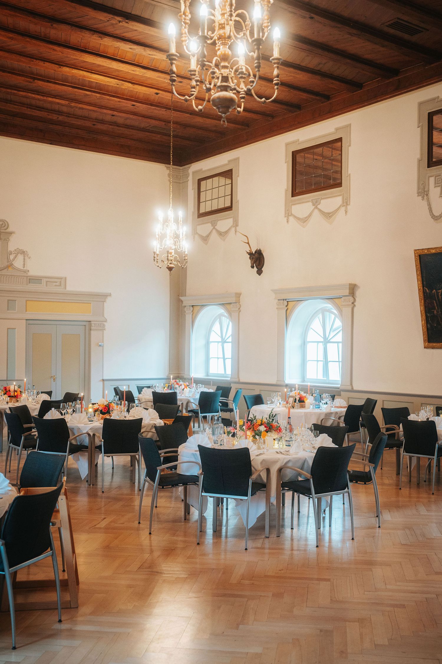Elegant historic banquet hall with wooden beam ceiling, crystal chandeliers, arched windows and round tables set for a wedding reception.