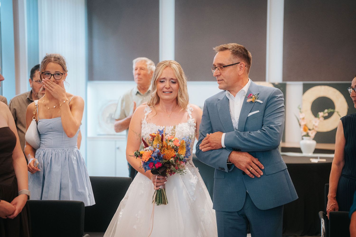 Smiling bride in a white lace gown holding a colorful bouquet walks down the aisle with a man in a blue suit as a guest tears up.