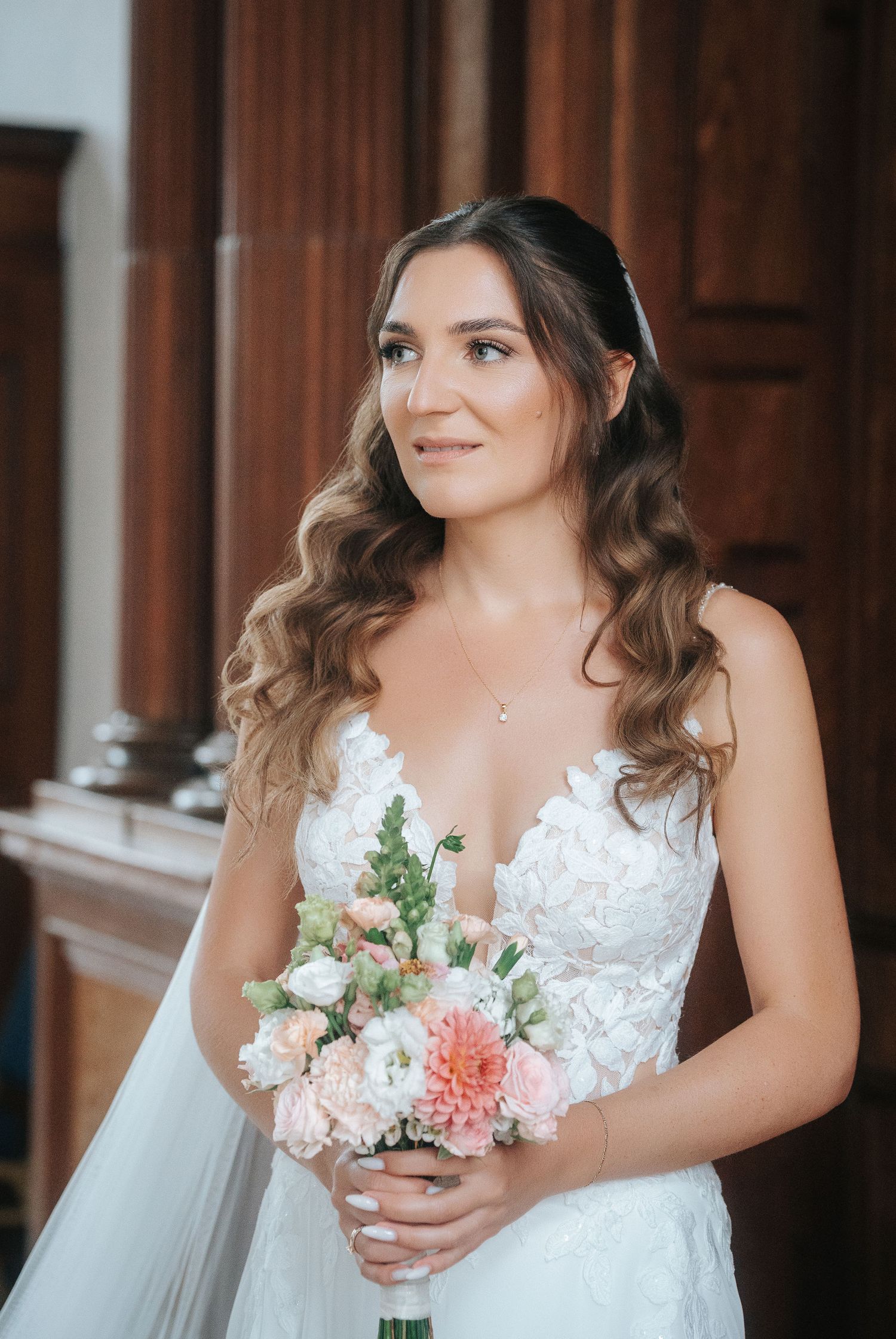 Smiling bride in a lace wedding dress with long wavy hair holding a pink and peach floral bouquet beside dark wooden paneling.