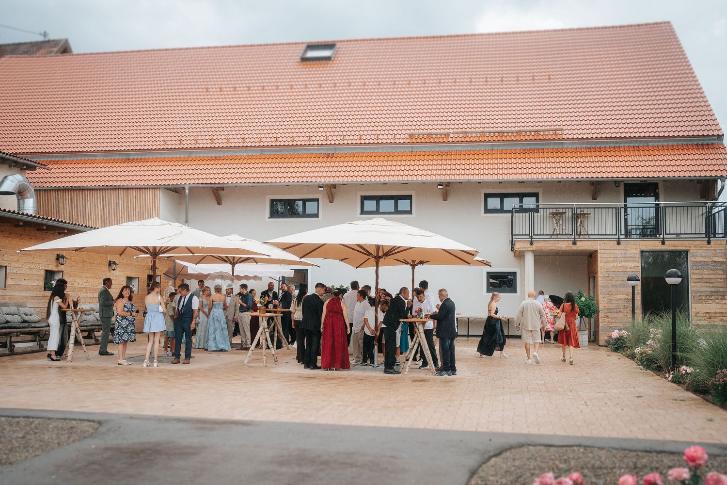 Wedding guests in colorful attire gather under white umbrellas on a stone patio outside a rustic venue with a terracotta tile roof.