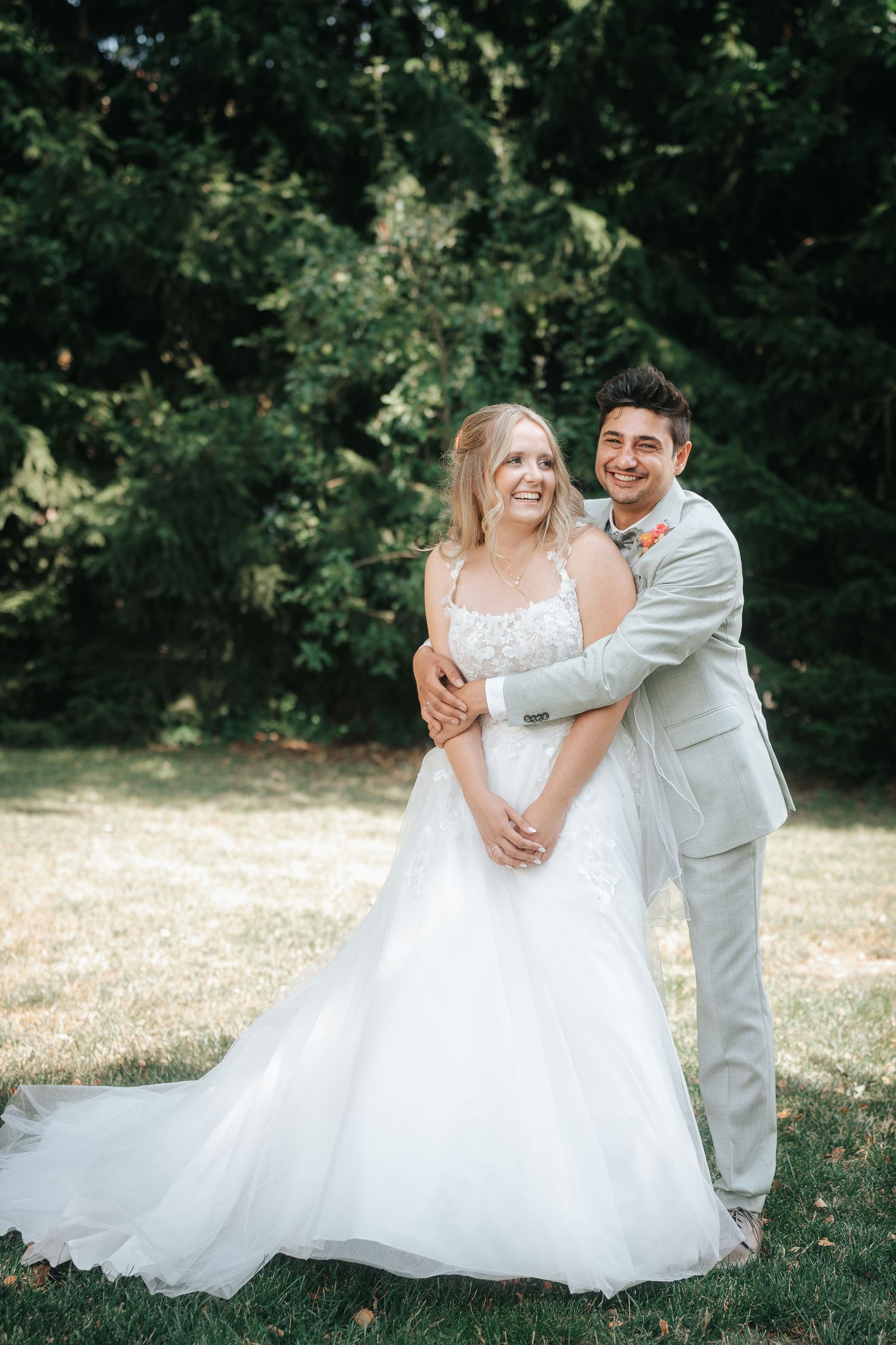 Smiling bride in a white lace wedding gown embraced from behind by a groom in a light gray suit on a sunny lawn with green trees.