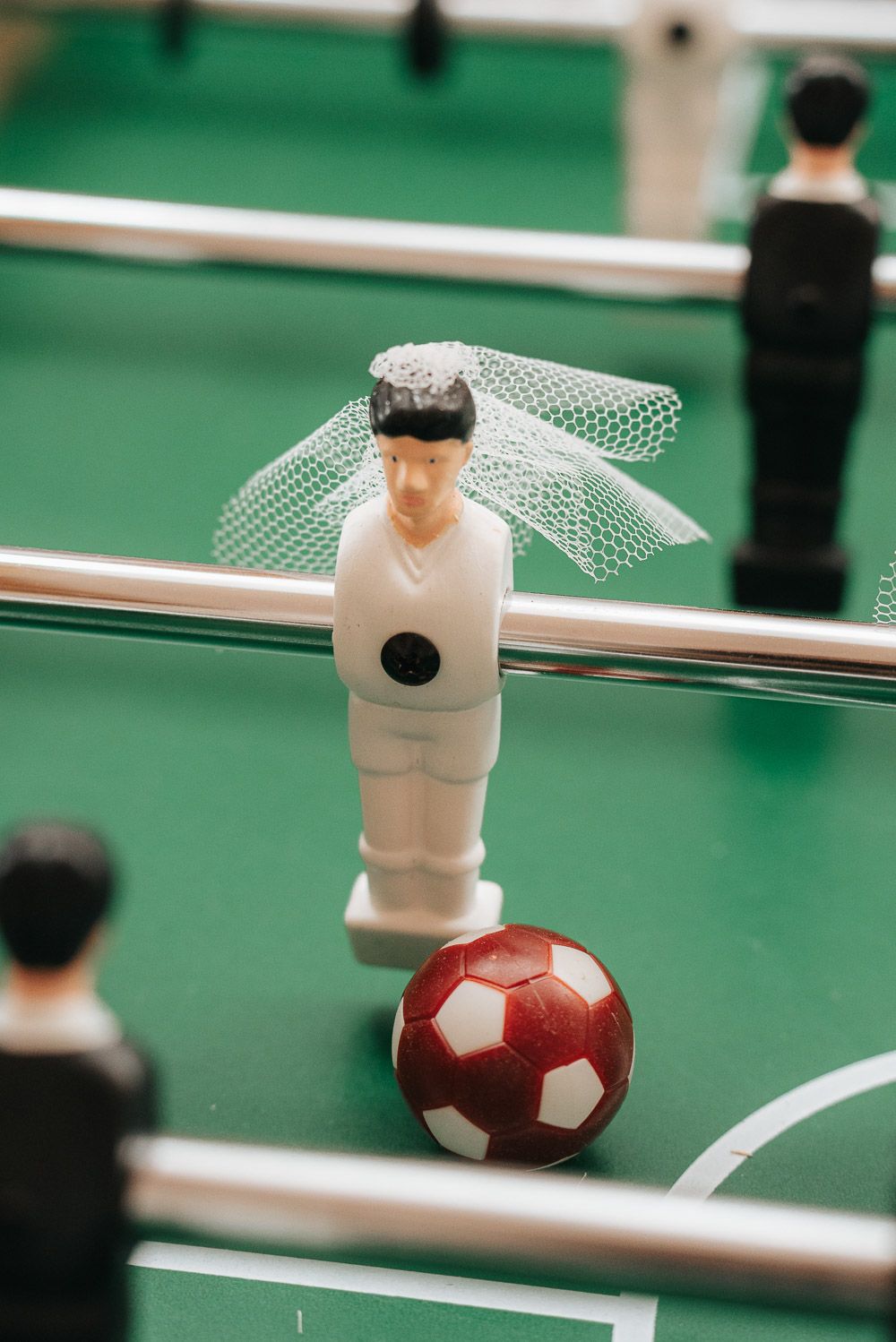 Close-up of a foosball table player figure wearing a bridal veil with a red and white ball on green playing field.