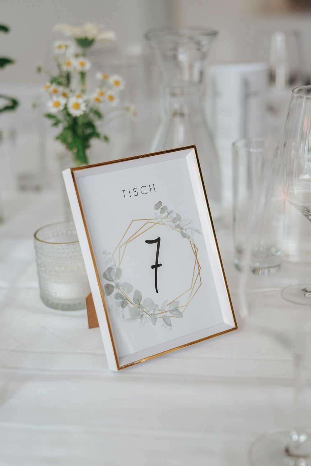 White wedding table setting with a floral table number card displaying number 7 and delicate daisies in the background.