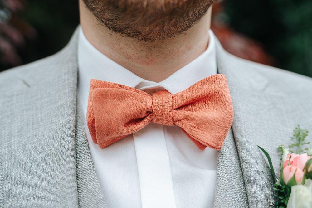 A person wearing a light gray suit and coral peach bow tie poses for a formal photo in an outdoor setting.