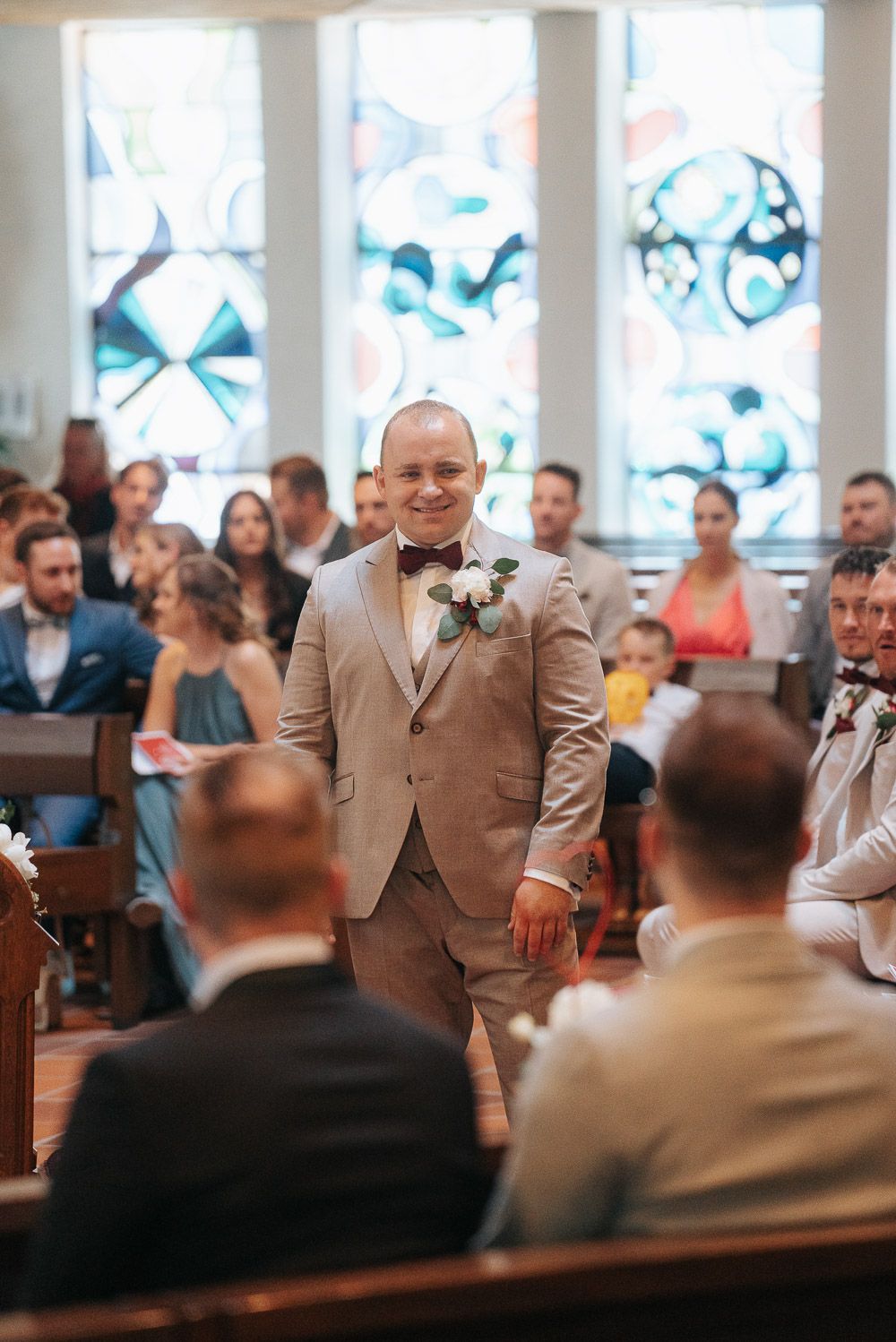 Wedding party in beige suits stand at the front of a church during a ceremony as guests look on from pews.