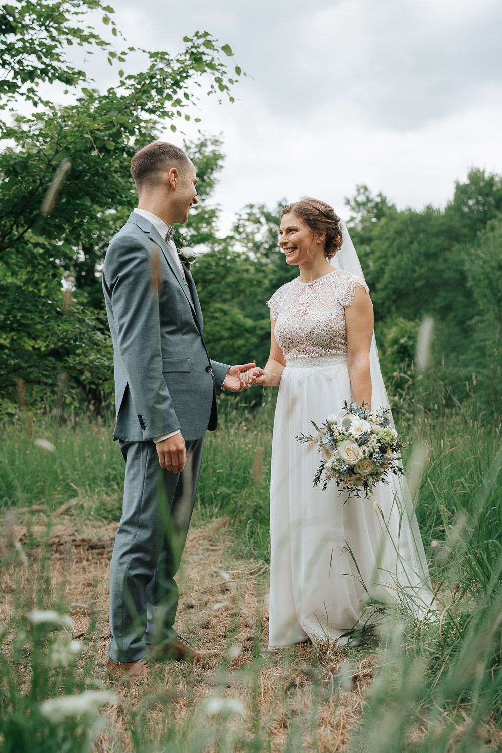 A couple in wedding attire shares a candid moment together in a scenic meadow surrounded by tall grass and greenery.