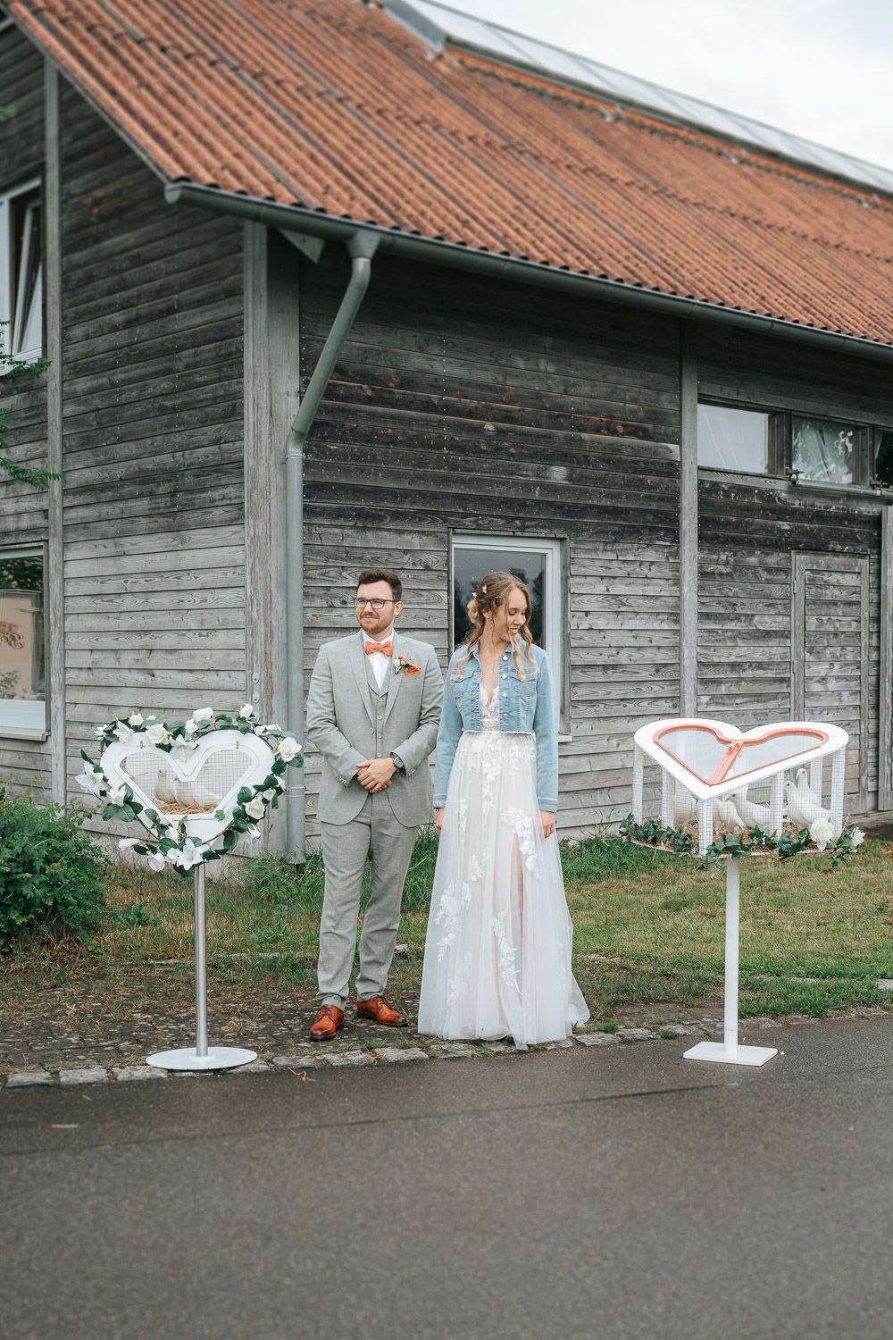 Wedding couple stands in front of rustic wooden barn exterior with autumn decorations and white drapery details.