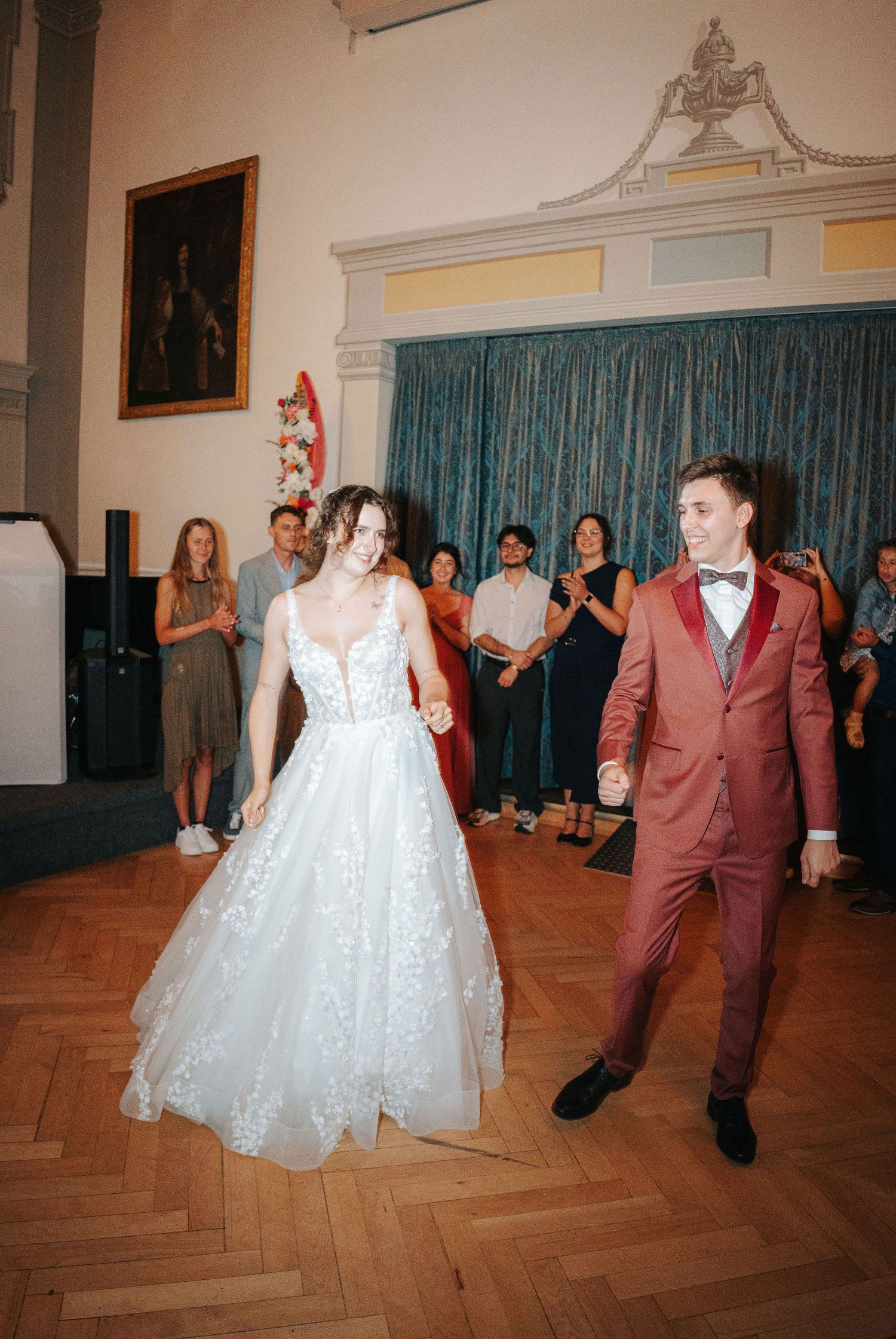 Bride in a lace gown and groom in a rust suit with bow tie share a first dance on a wood floor as wedding guests watch.