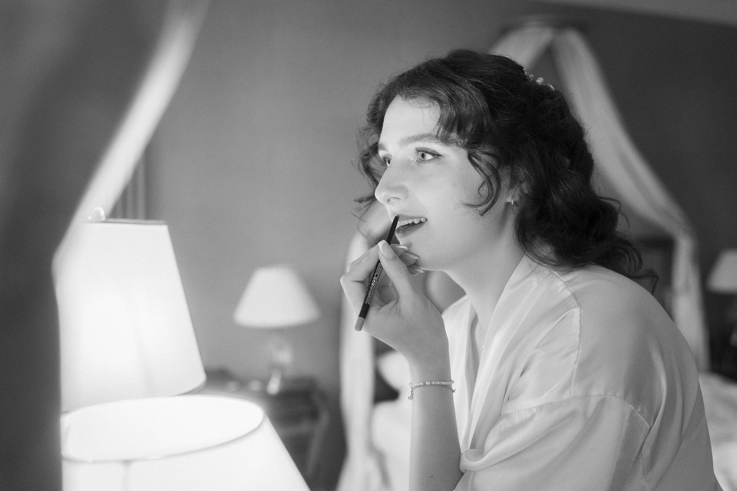Black and white photo of a smiling woman with curly updo hair applying makeup in a softly lit bedroom beside a glowing lamp.