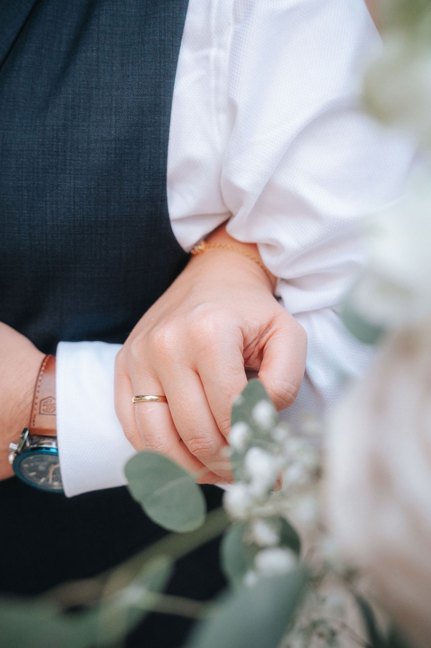 Close-up of a groom's hand with a gold wedding band, navy vest, white shirt cuff and wristwatch, framed by white flowers and eucalyptus.