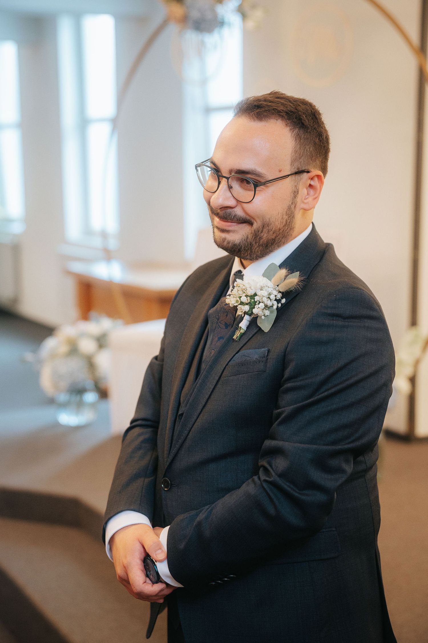 Smiling groom in a dark pinstripe suit and glasses wearing a white floral boutonniere stands at a wedding ceremony.
