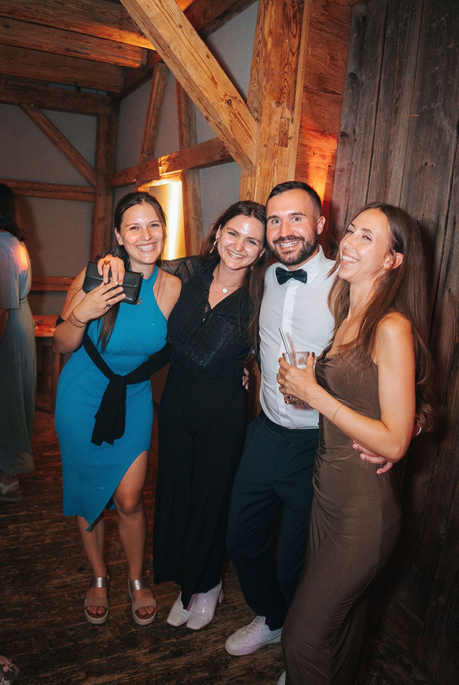 Four smiling guests pose arm in arm at a festive party inside a rustic wooden barn with warm lighting, three women and a man in a bow tie.