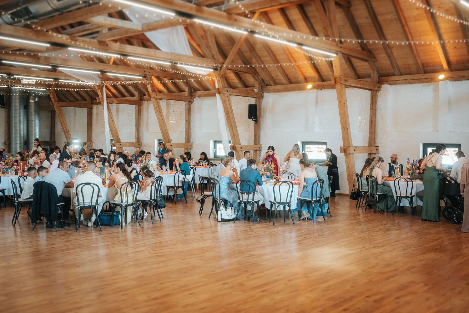 Wedding guests seated at round tables in a rustic barn venue with exposed wooden beams, string lights and a polished wood dance floor.