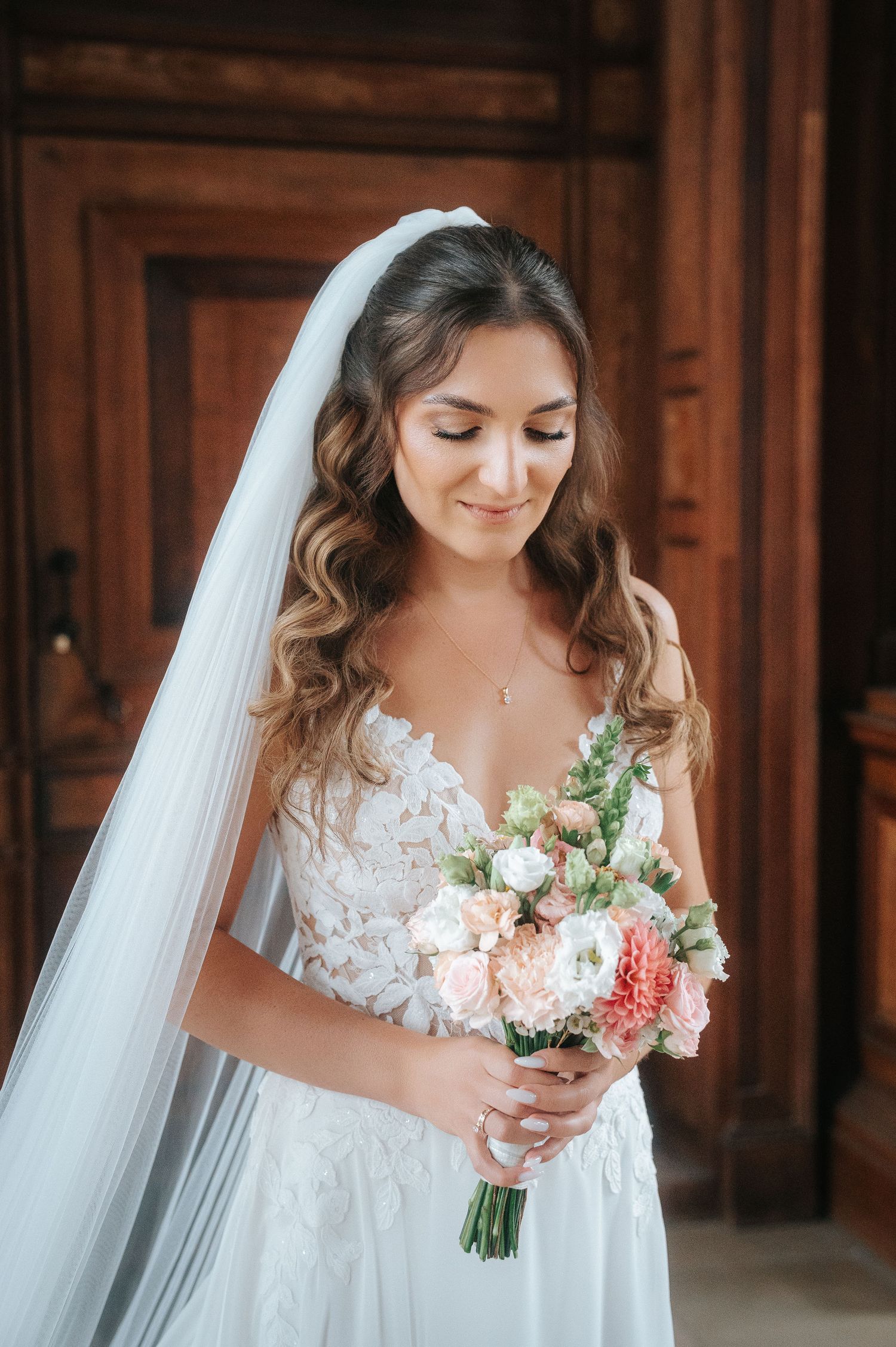 Smiling bride in a lace wedding gown and long veil holds a pink and white floral bouquet in front of a carved wooden door.