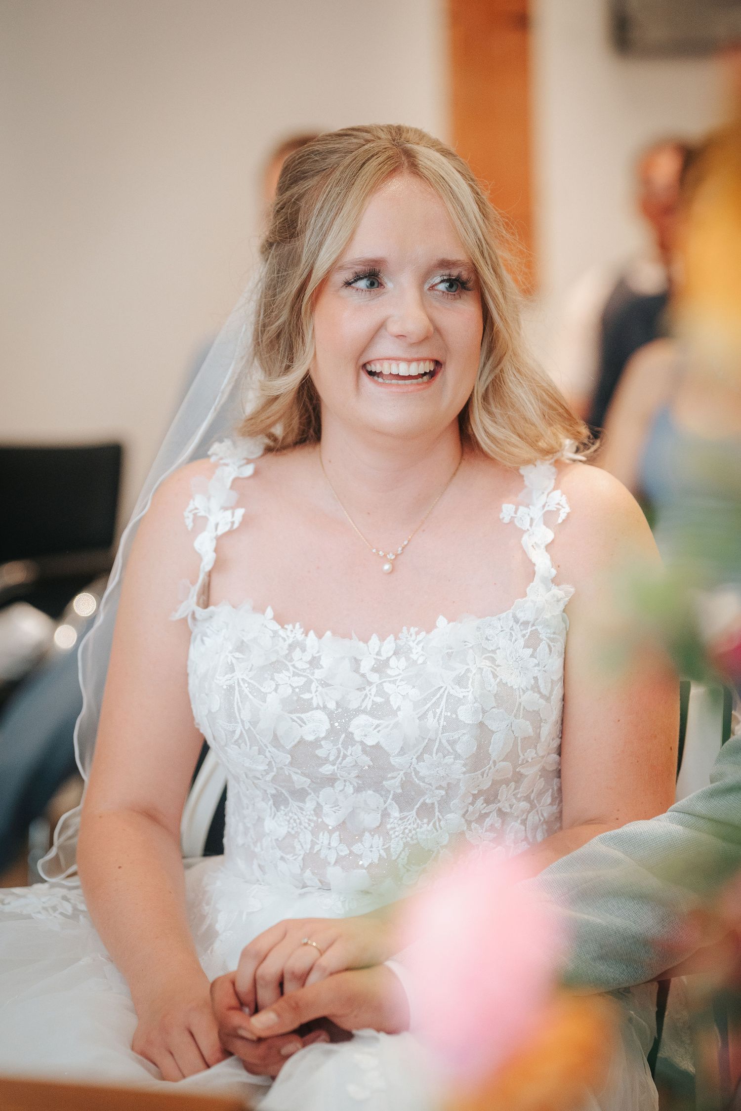 Smiling bride in a white lace wedding dress and veil holds hands with her partner during an indoor wedding ceremony.