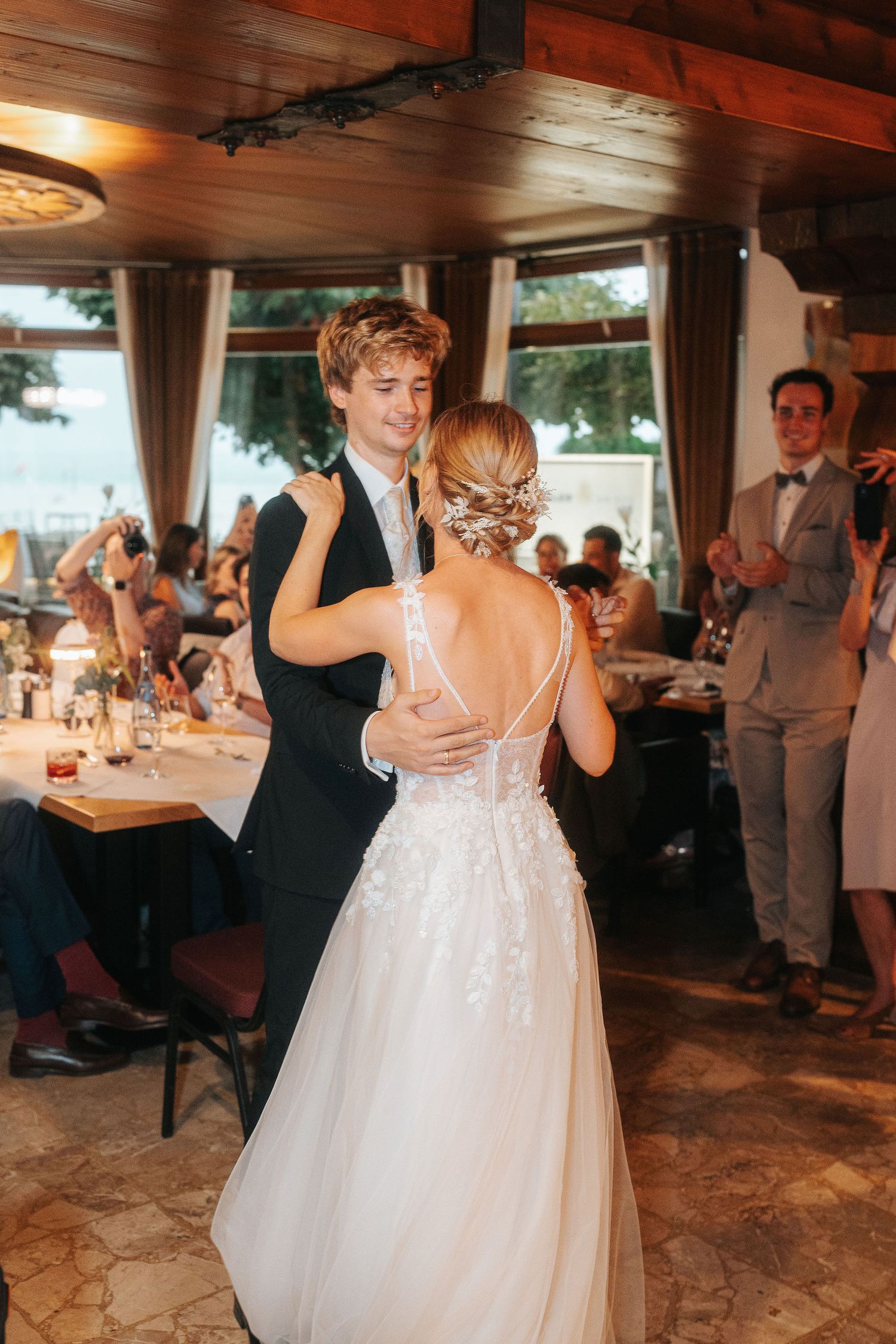 Bride in a lace wedding gown and groom in a dark suit share a first dance as guests watch and applaud at a reception.