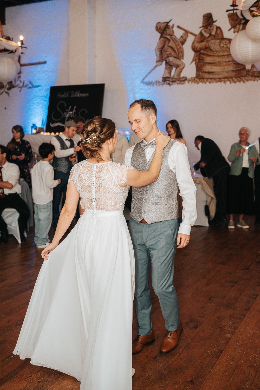 Couple shares their first dance at wedding reception with rustic wall art and hardwood floor in background.