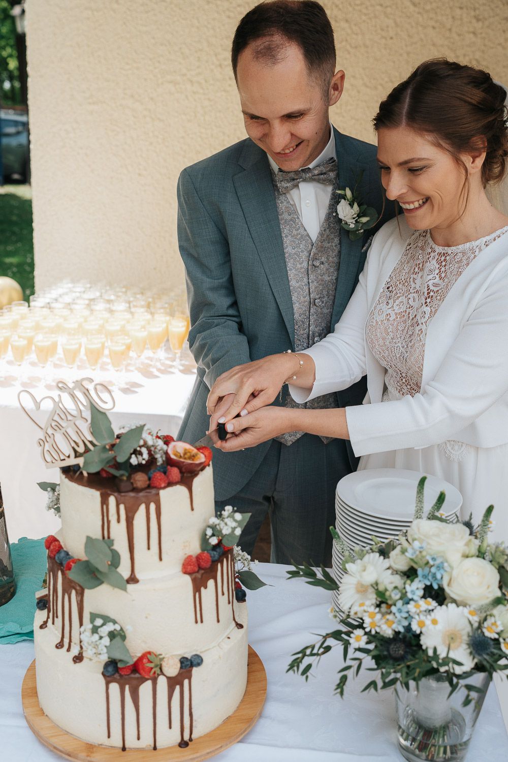 Cutting a white wedding cake with chocolate drips, fresh flowers and eucalyptus, with a floral bouquet on the table.