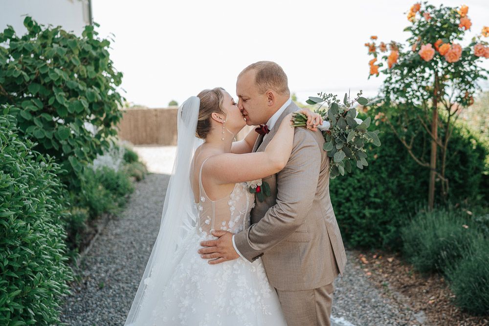 A couple shares their first kiss as newlyweds on a garden path surrounded by lush greenery and orange flowers.