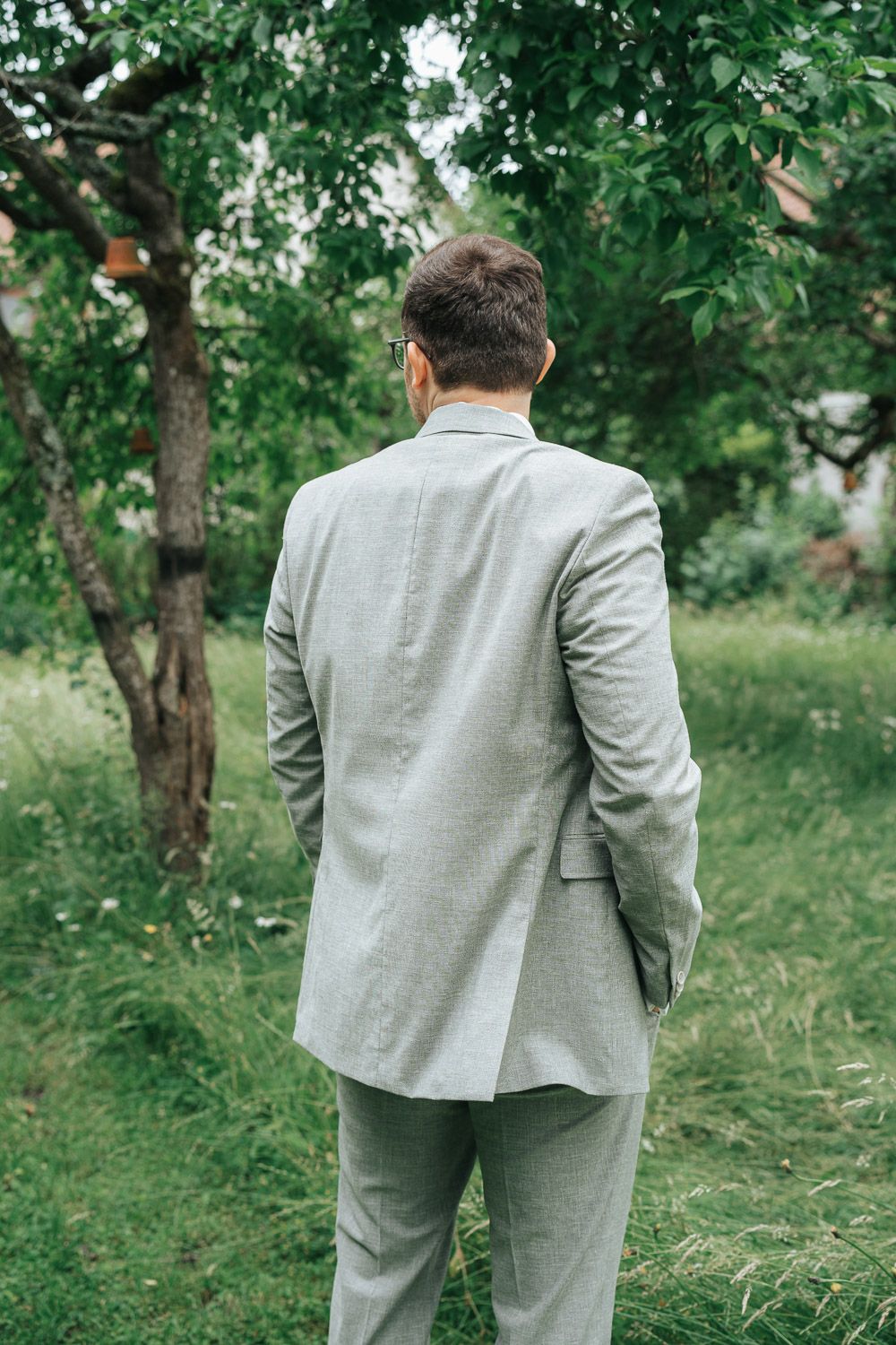View from behind of someone wearing a light gray suit standing in a lush garden setting with green trees.