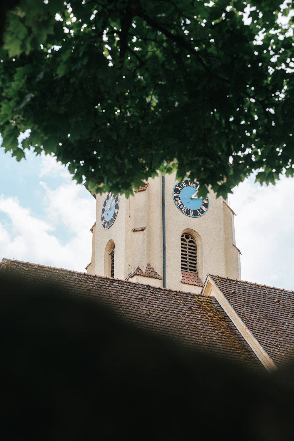 Church steeple with a blue clock face rises above a brown tiled roof, framed by green summer leaves overhead.