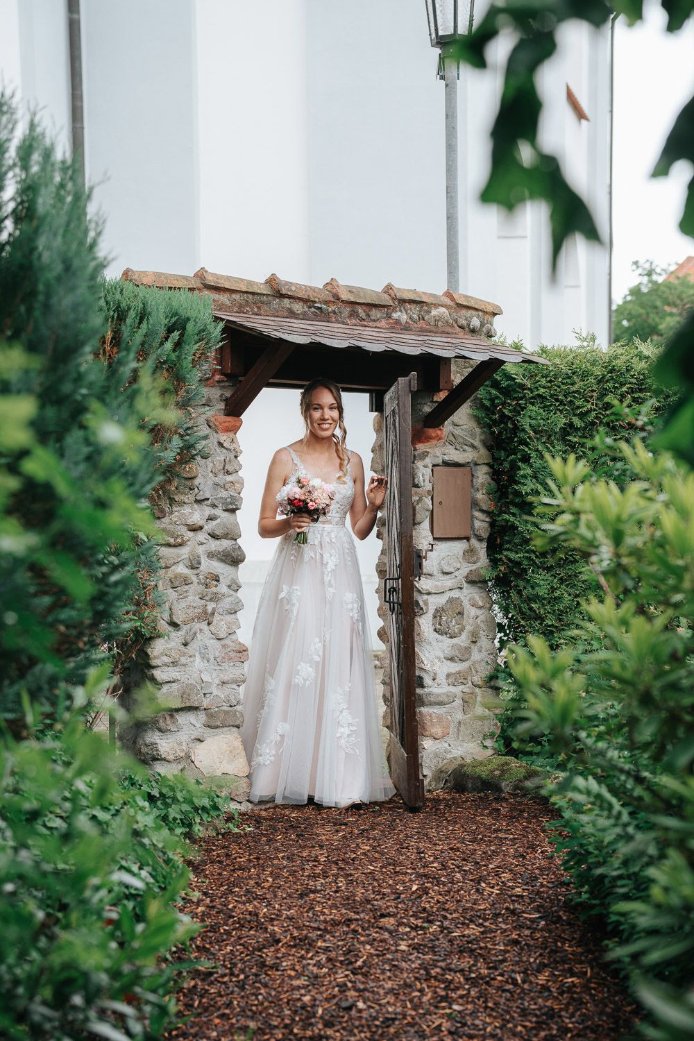 A bride poses in an elegant white dress beneath a stone archway along a mulch pathway framed by greenery.
