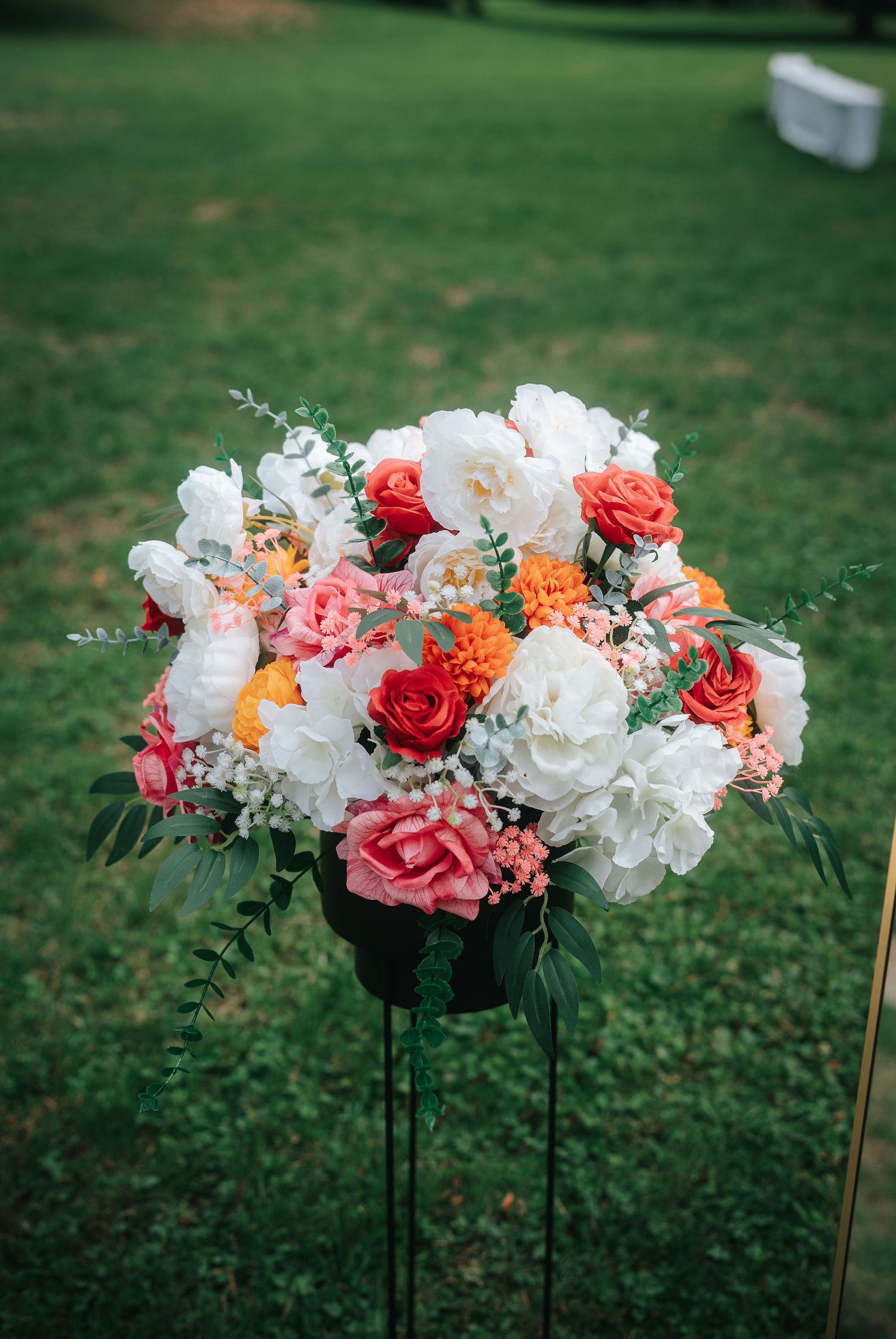 Wedding floral arrangement of white, coral, orange and pink roses with greenery on a tall black stand in a sunny grassy lawn.