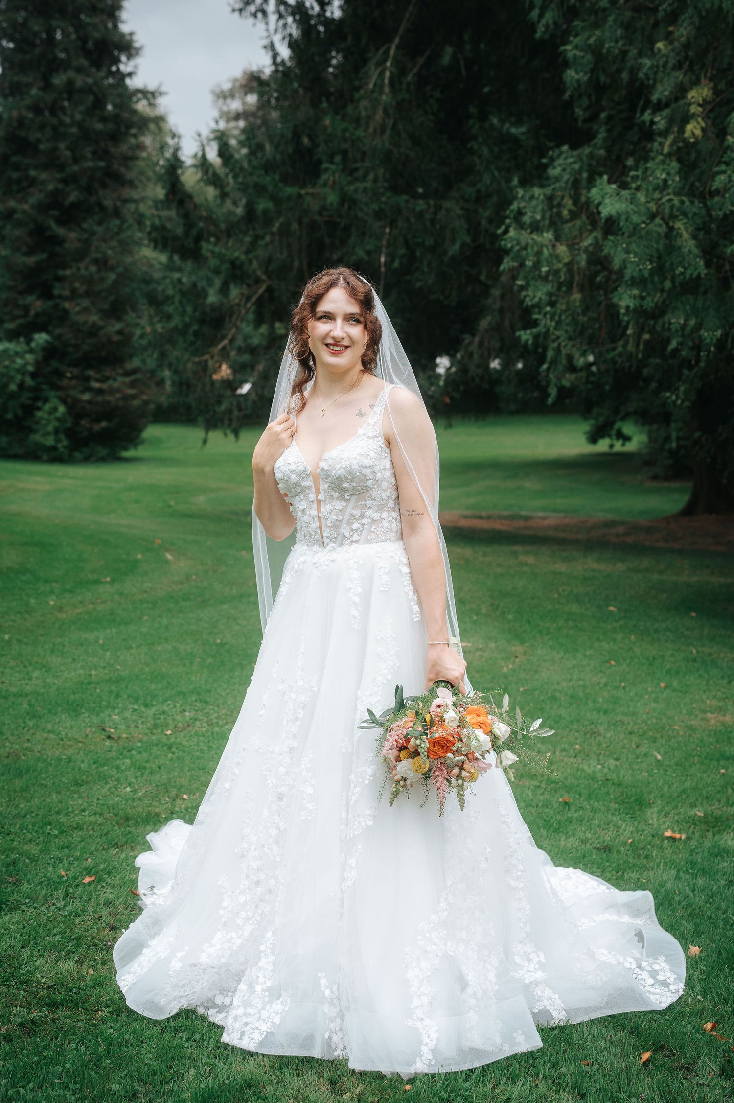 Smiling bride in a white lace wedding gown and veil holds an orange and peach floral bouquet on a green lawn surrounded by tall trees.
