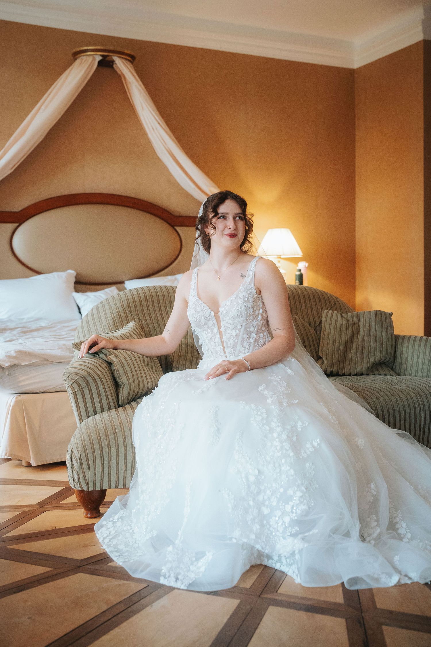 A bride in a white lace wedding gown sits on a striped sofa in an elegant hotel suite with a canopy bed and warm lamplight.