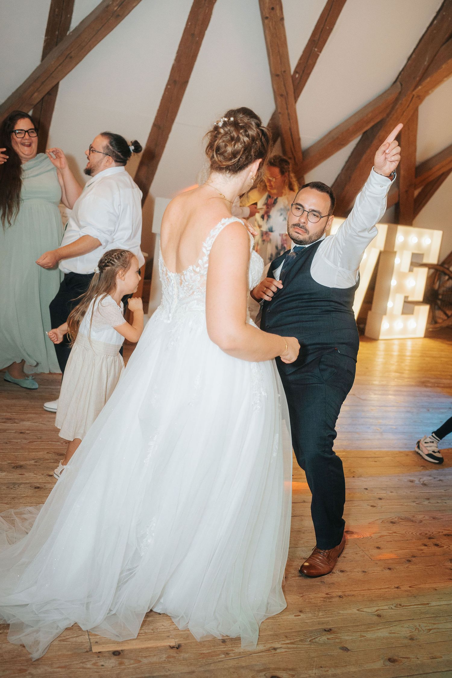 Bride in a white lace gown dances with a groom in a vest on a wooden floor while guests and a flower girl celebrate under rustic beams.