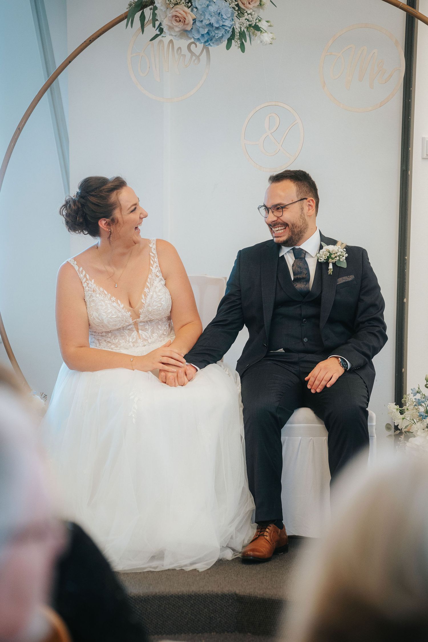 Smiling bride in a lace-bodice wedding gown holds hands with her bearded groom in a navy suit during a ceremony beside a floral hoop arch.