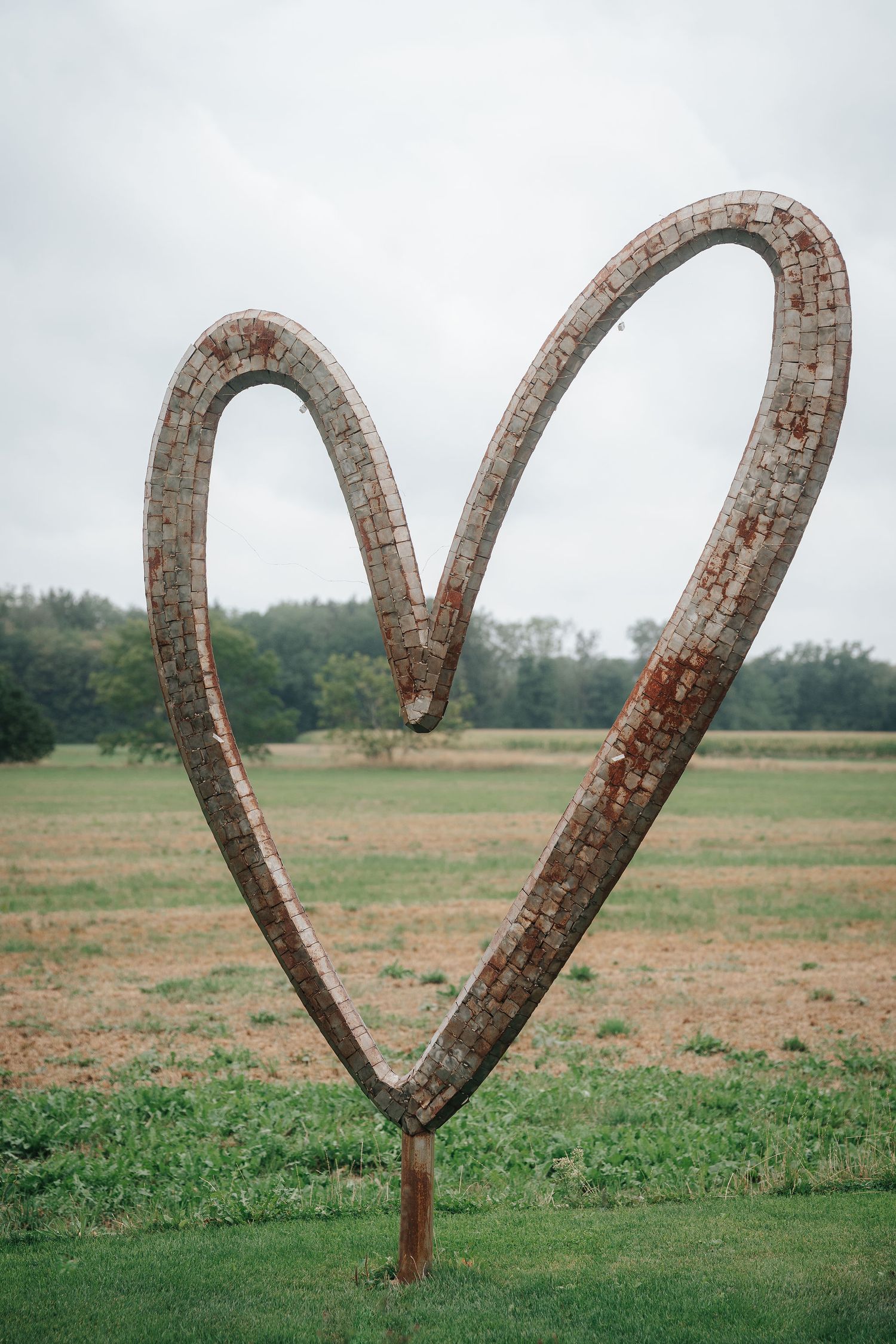Large rusted heart-shaped metal sculpture standing in a grassy field with trees in the background under an overcast sky.