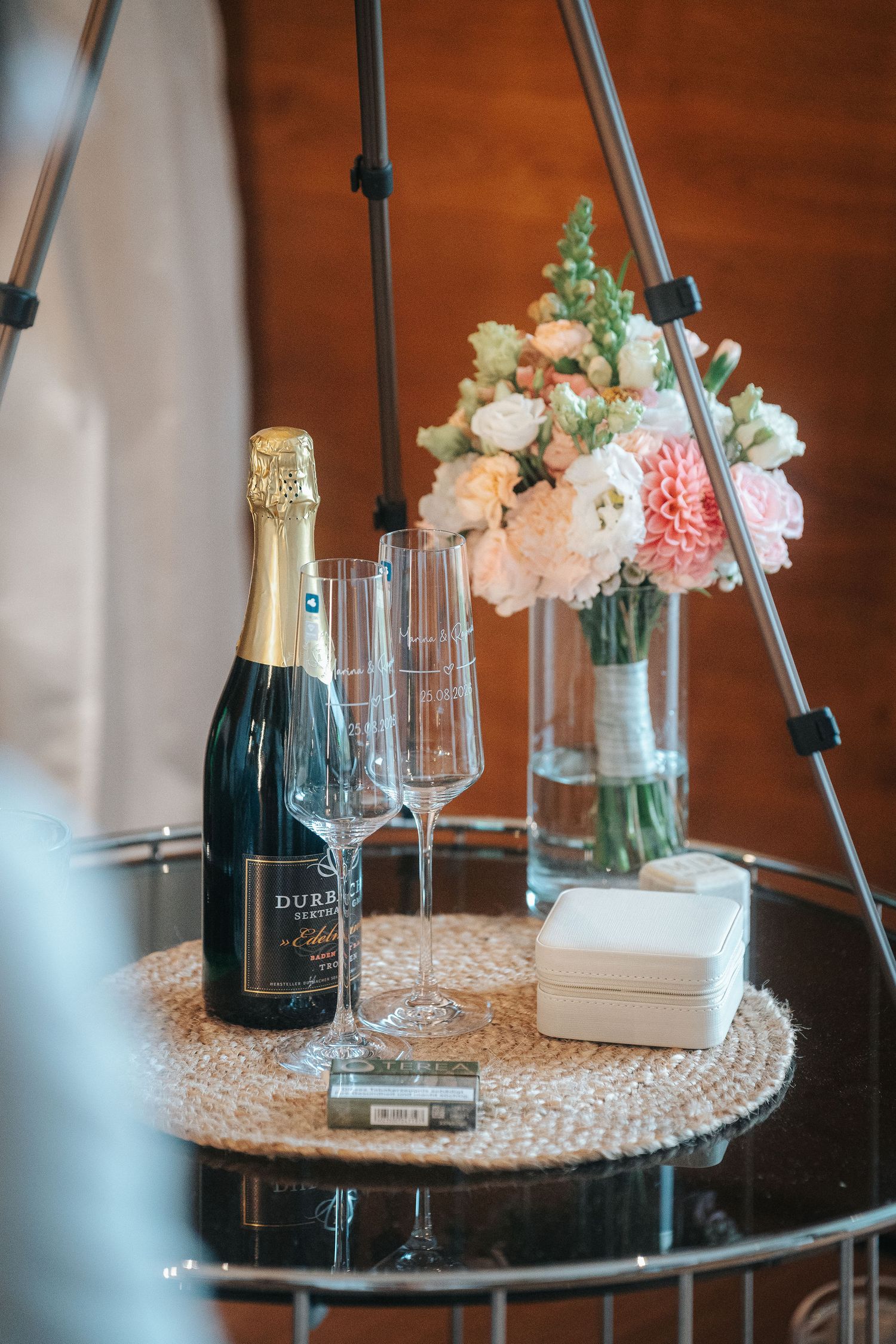 Champagne bottle, two flutes, a white keepsake box and a pink and white flower bouquet on a woven placemat atop a round glass table.