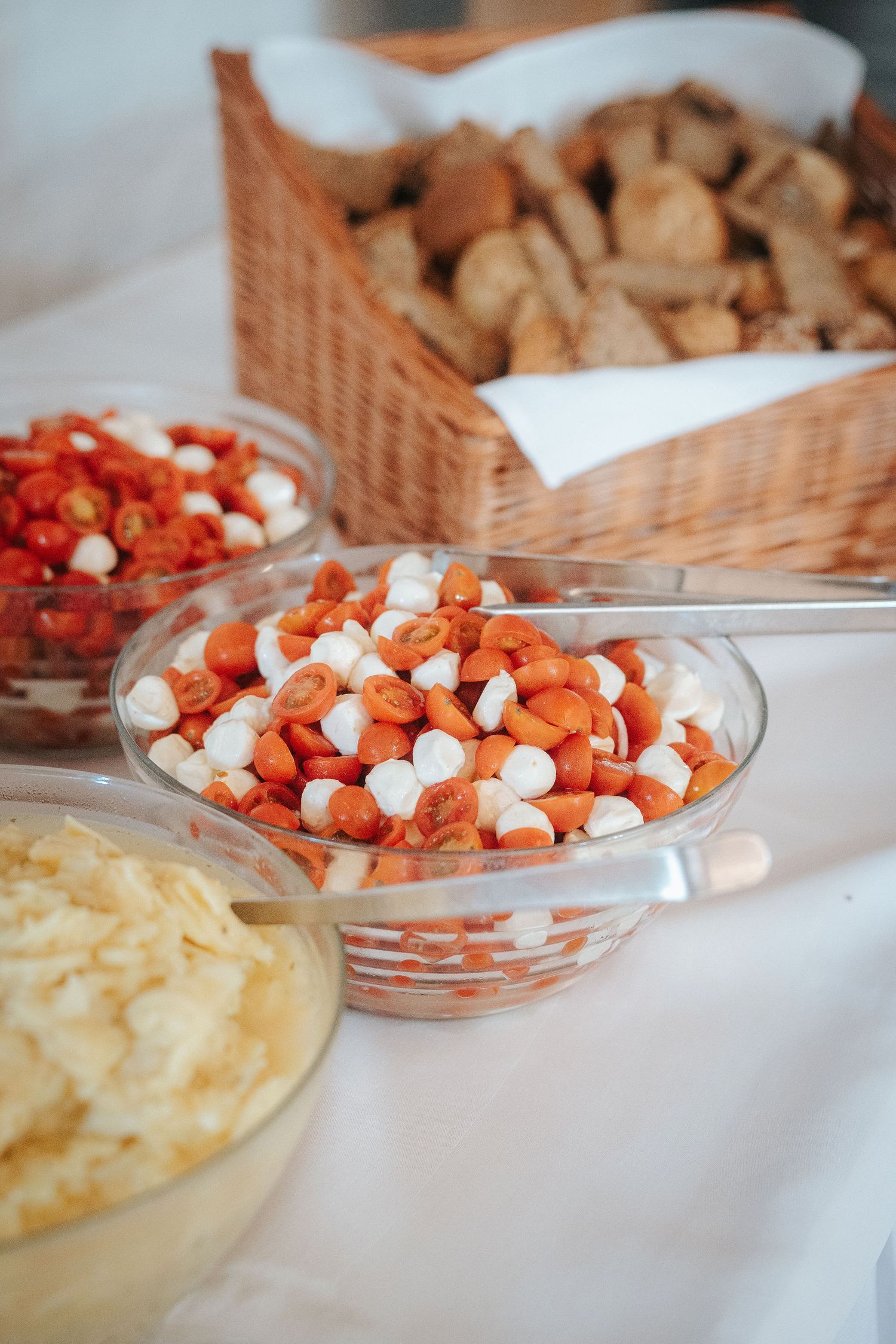 Glass bowls of cherry tomatoes and mozzarella pearls on a buffet table beside a wicker basket of rustic bread and a bowl of shredded cheese.
