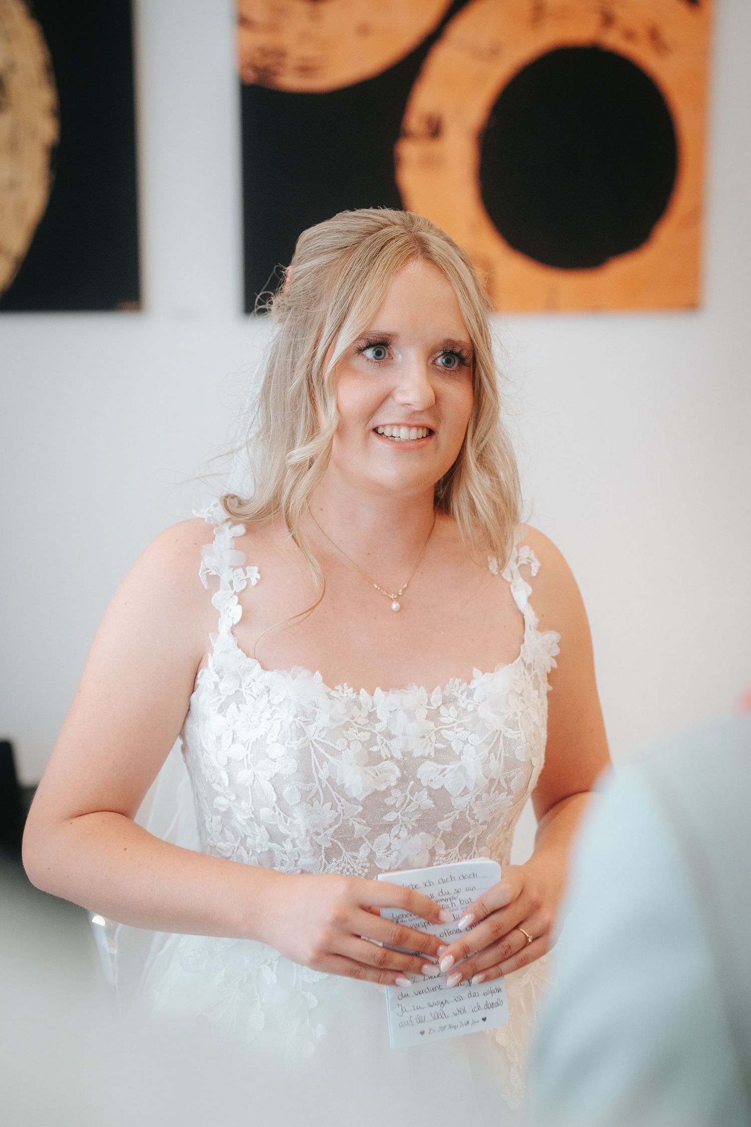 Bride in a white lace wedding gown with a delicate necklace smiles while holding a small card during the ceremony.