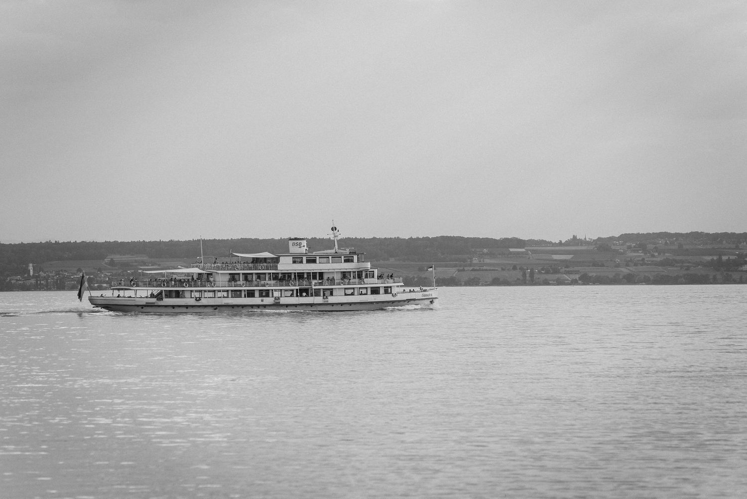 Black and white photo of a classic paddle steamer ferry boat cruising on calm lake waters with hilly shoreline in background.