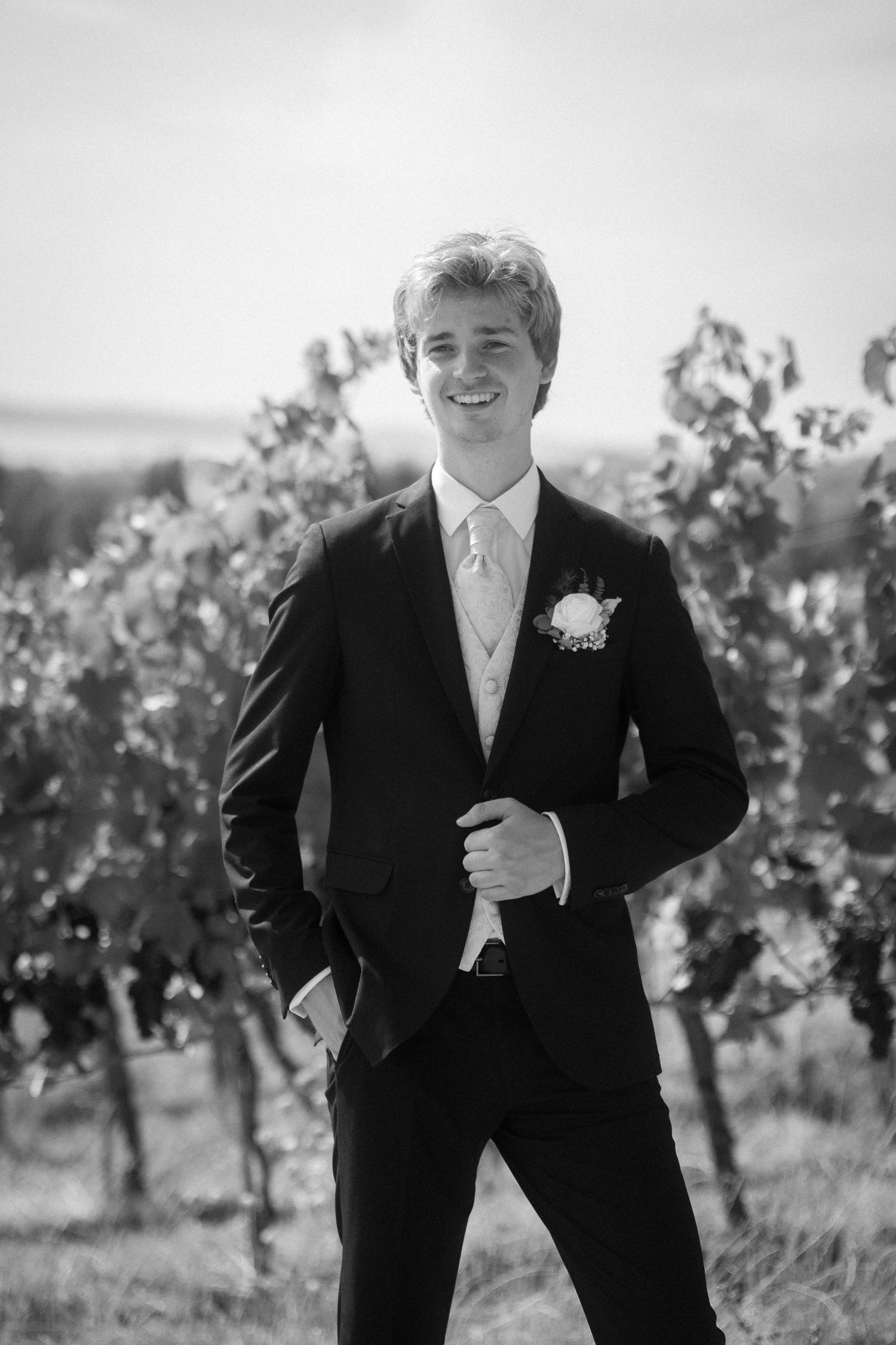 Black and white portrait of a smiling groom in a dark suit with a boutonniere standing among grapevines on his wedding day.