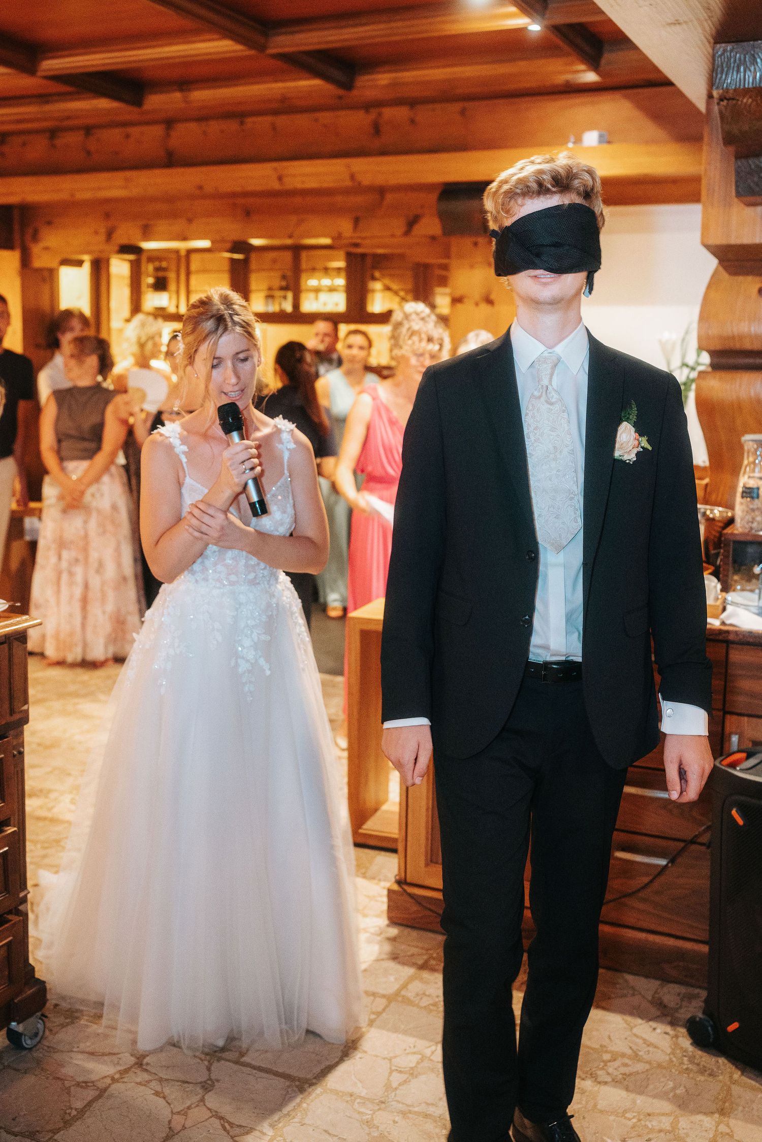 Bride in a white tulle gown speaks into a microphone beside a blindfolded groom in a black suit during a wedding game as guests watch.
