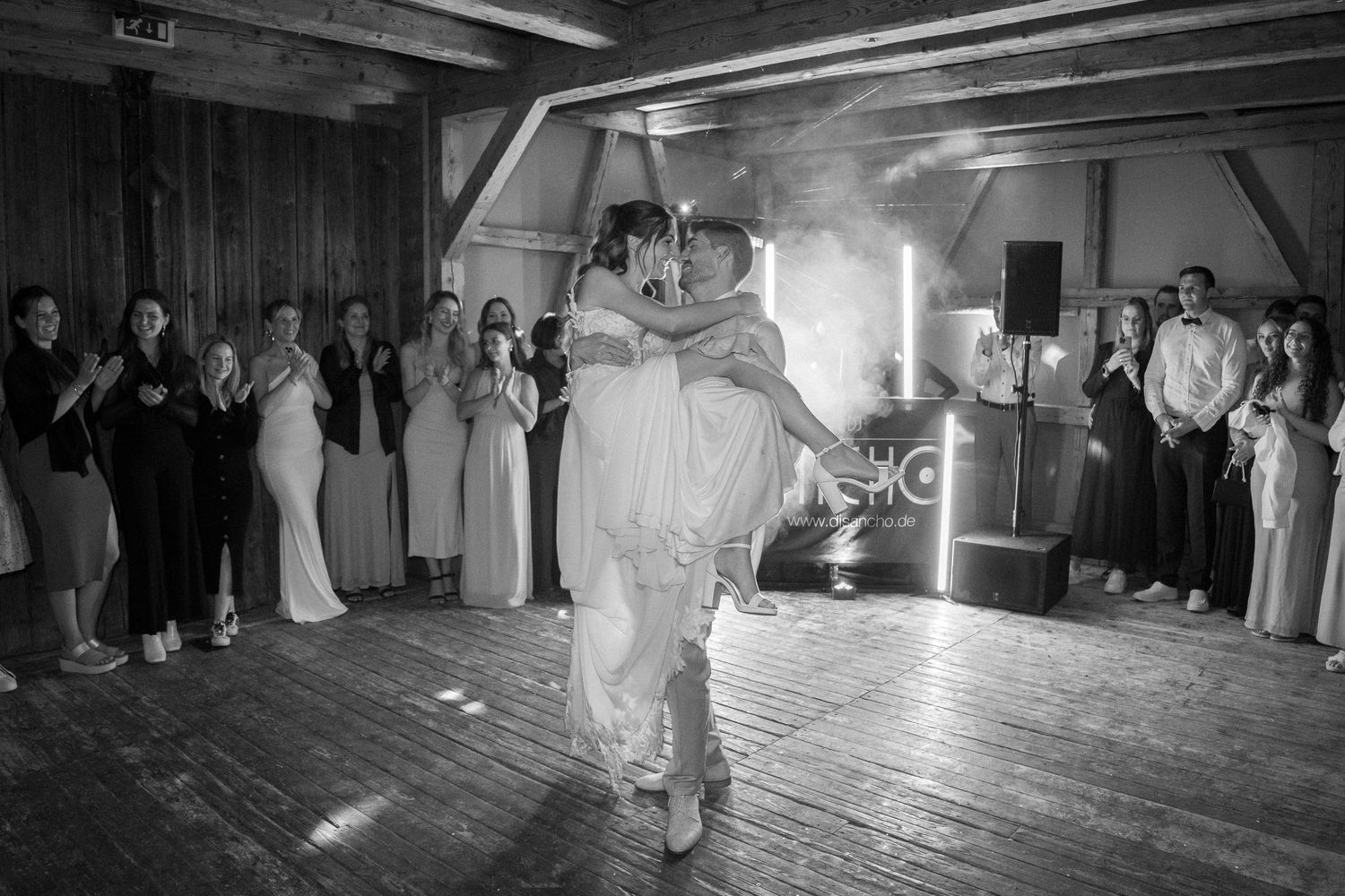 Black and white photo of groom lifting bride during first dance at wedding reception with guests watching.
