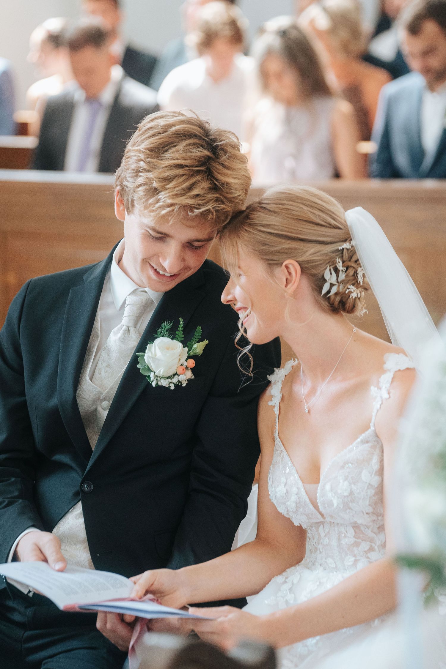 A smiling bride and groom share a joyful moment reading a wedding program together during their church ceremony.