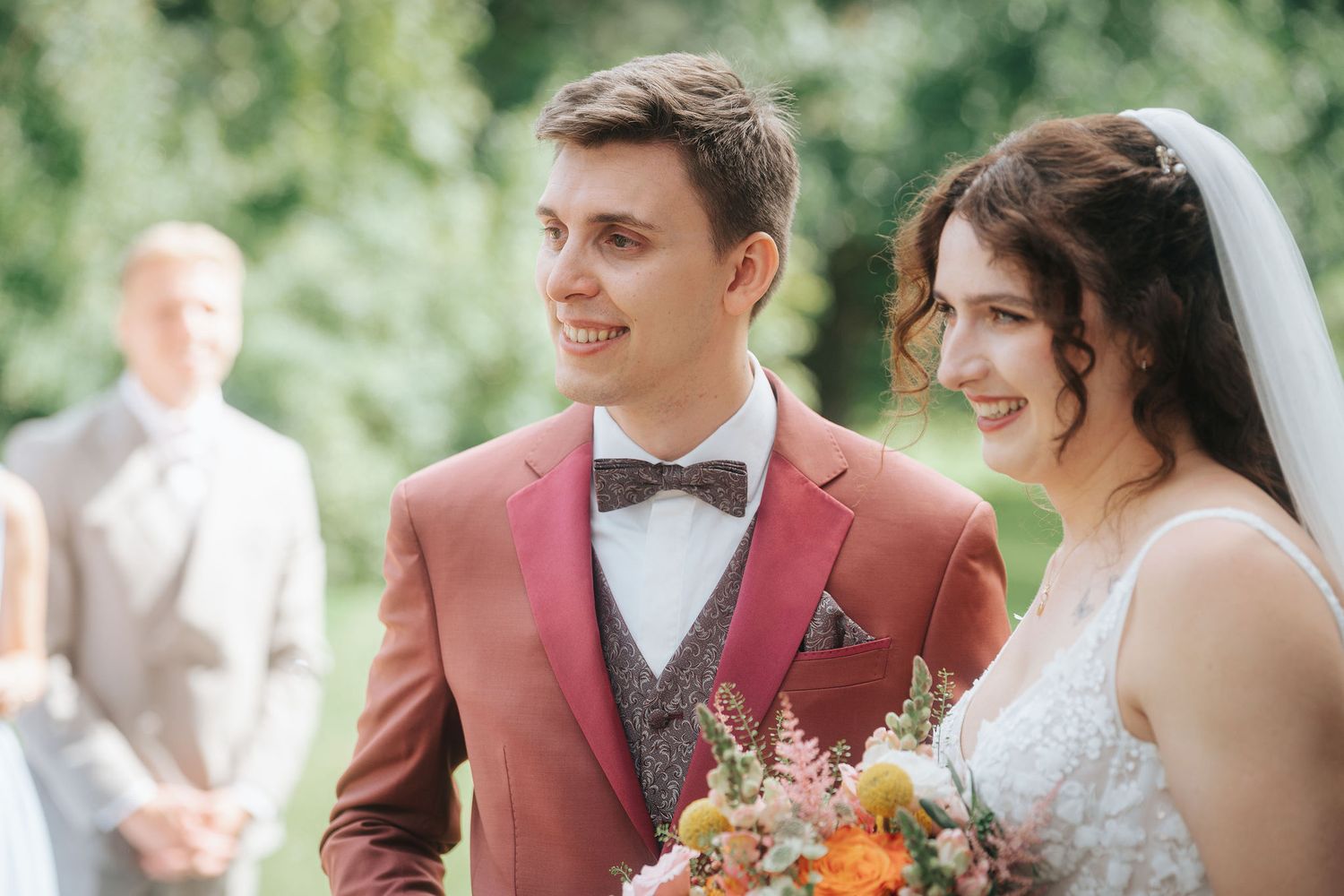 Smiling groom in a rust-colored suit and bow tie stands beside a bride in a white dress and veil holding a colorful bouquet outdoors.