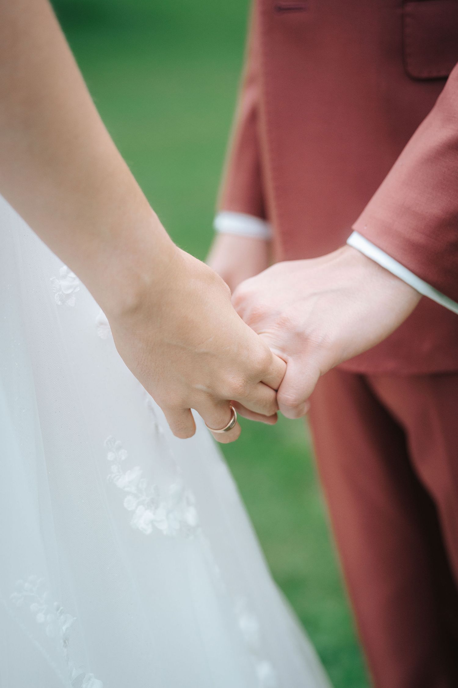 Bride in a white lace gown holds hands with a groom in a rust-colored suit on a green lawn, her gold wedding ring visible.