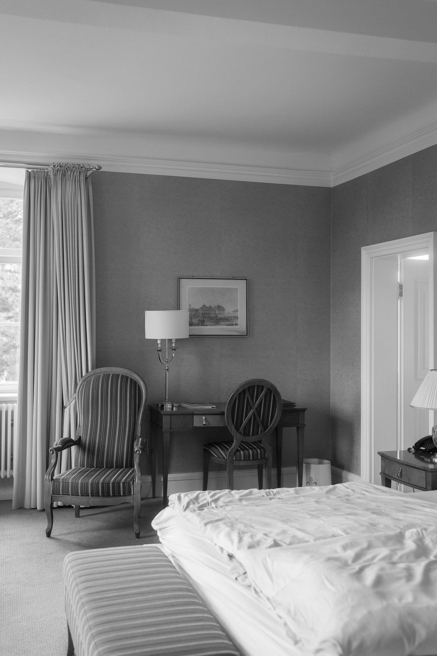Black and white photo of an elegant hotel bedroom with a striped armchair, writing desk, framed artwork, lamp, and a rumpled bed.