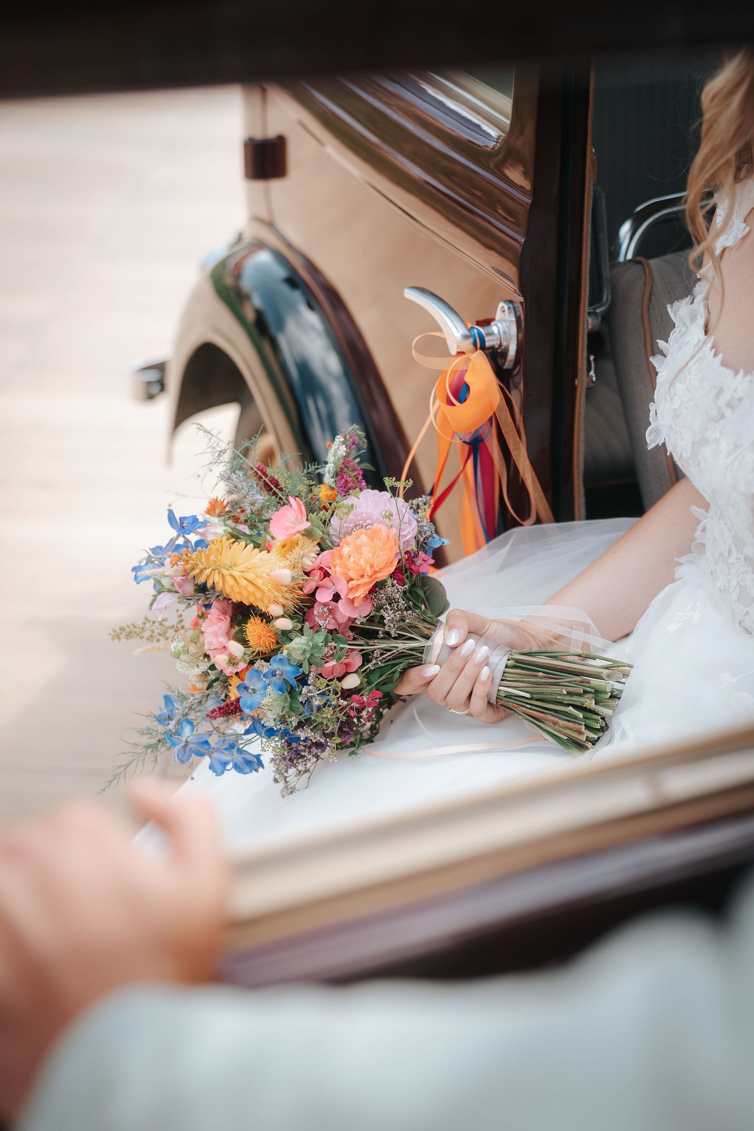 Bride in a lace gown holds a colorful wildflower bouquet with orange ribbons beside a vintage car as a hand reaches in.