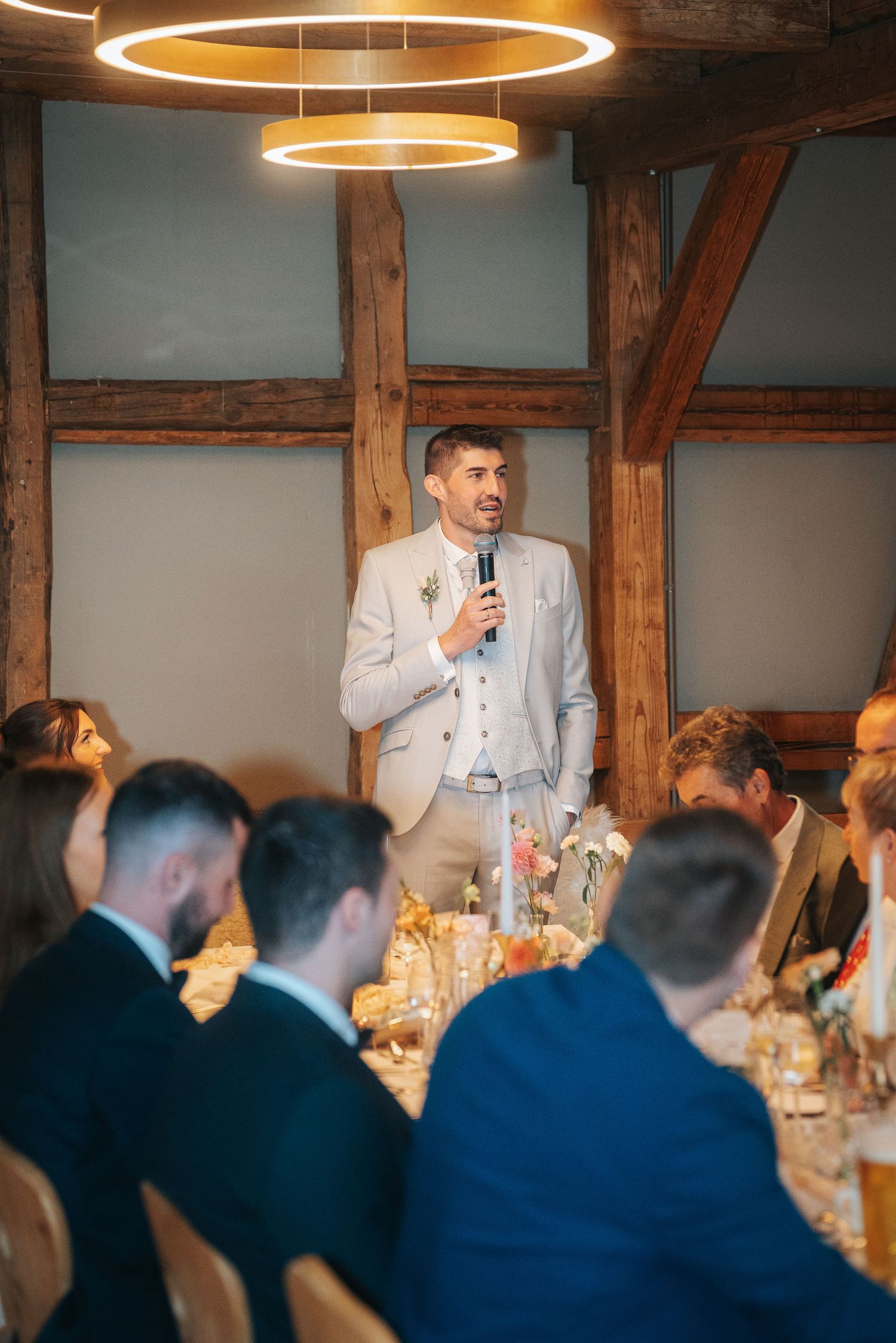 Man in a light suit holds a microphone and gives a speech to seated guests at a rustic wedding reception with timber beams.